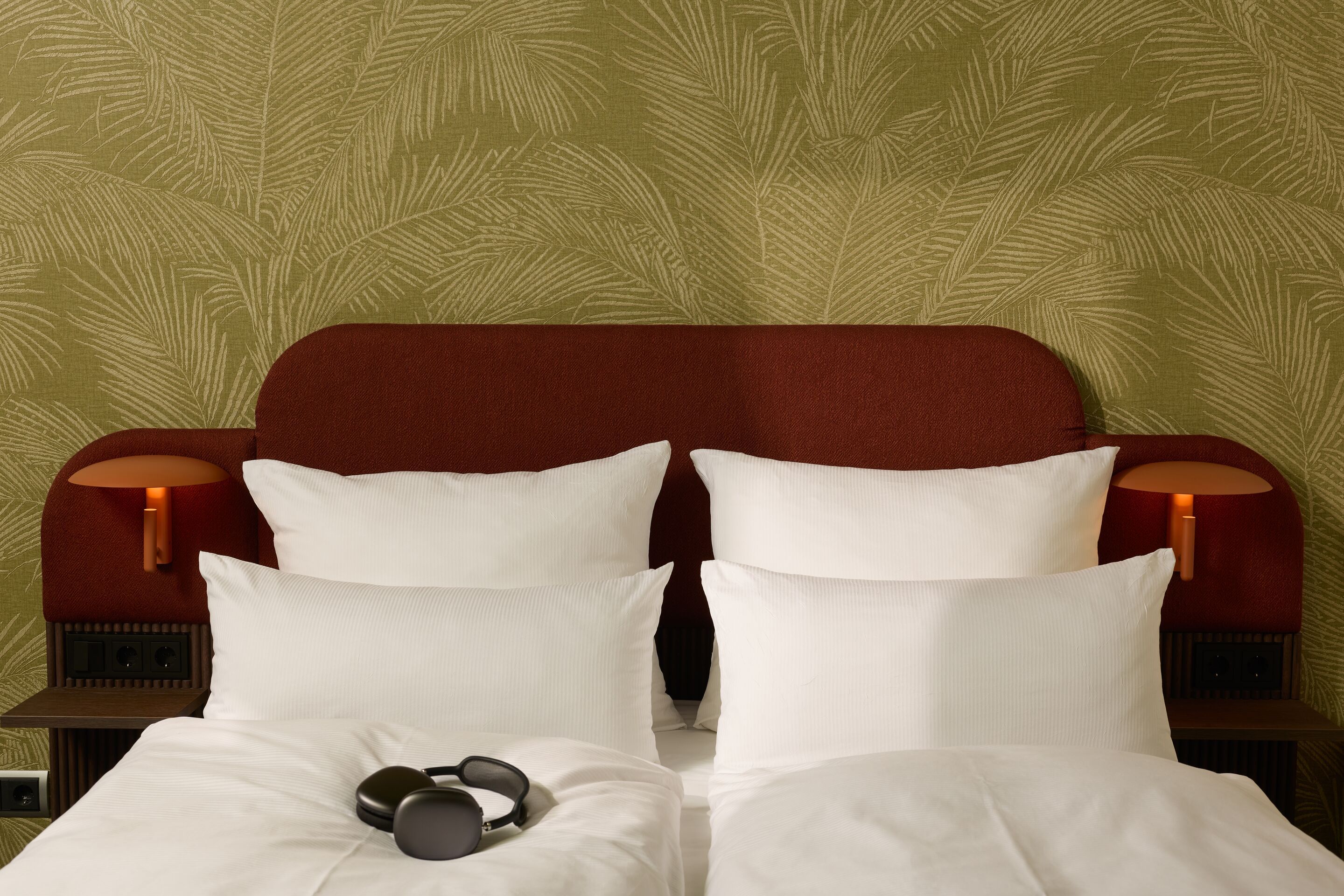 Hotel Frankfurt Metropolitan The Cloud One Doppelbett mit weißer Bettwäsche und weichen Kissen, grüne Tapete im Hintergrund, kabellose Kopfhörer auf der Bettdecke laden zum Entspannen ein.