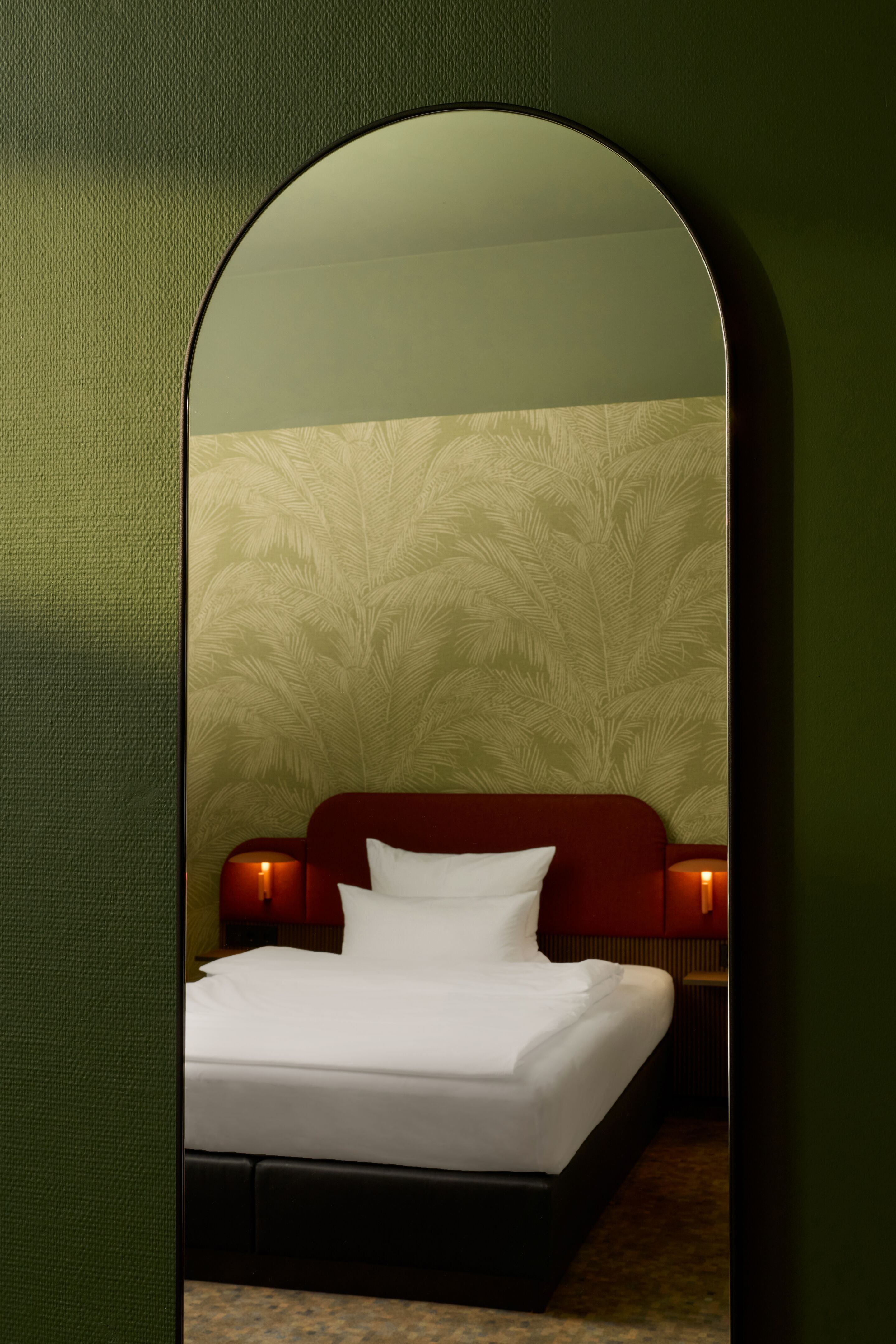 Hotel Frankfurt Metropolitan The Cloud One Blick durch einen runden Spiegel auf ein Hotelzimmer mit einem großen Bett, roten Kopfteil, weißen Kissen und dezent gemusterter Wand im Hintergrund. Zwei kleine Nachttischlampen sorgen für warmes, gemütliches Licht.