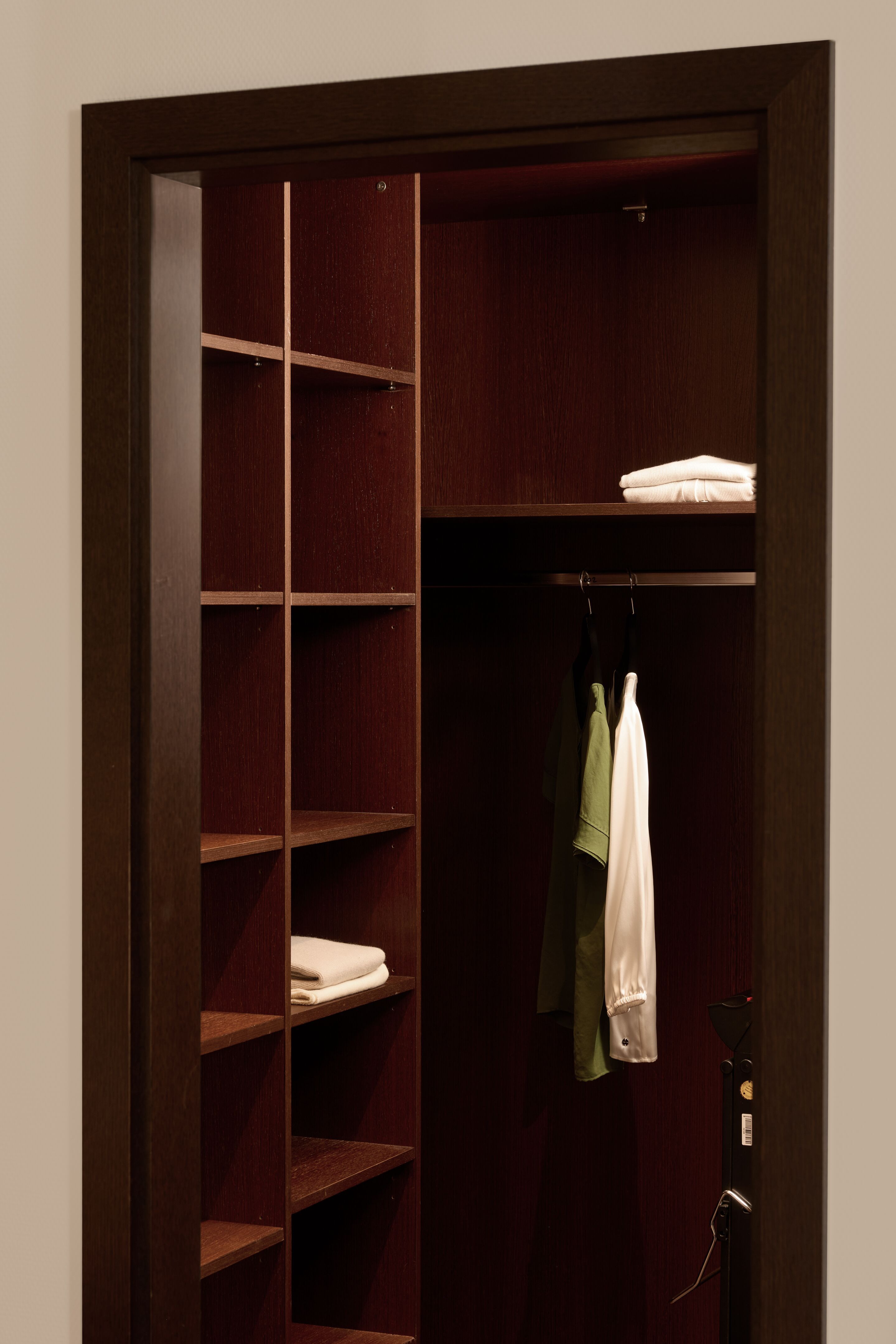 Hotel Frankfurt Metropolitan The Cloud One Zimmer mit offener, dunkler Holzschrank mit mehreren Regalfächern, in denen einige gefaltete Handtücher liegen, und zwei aufgehängten Kleidungsstücken.