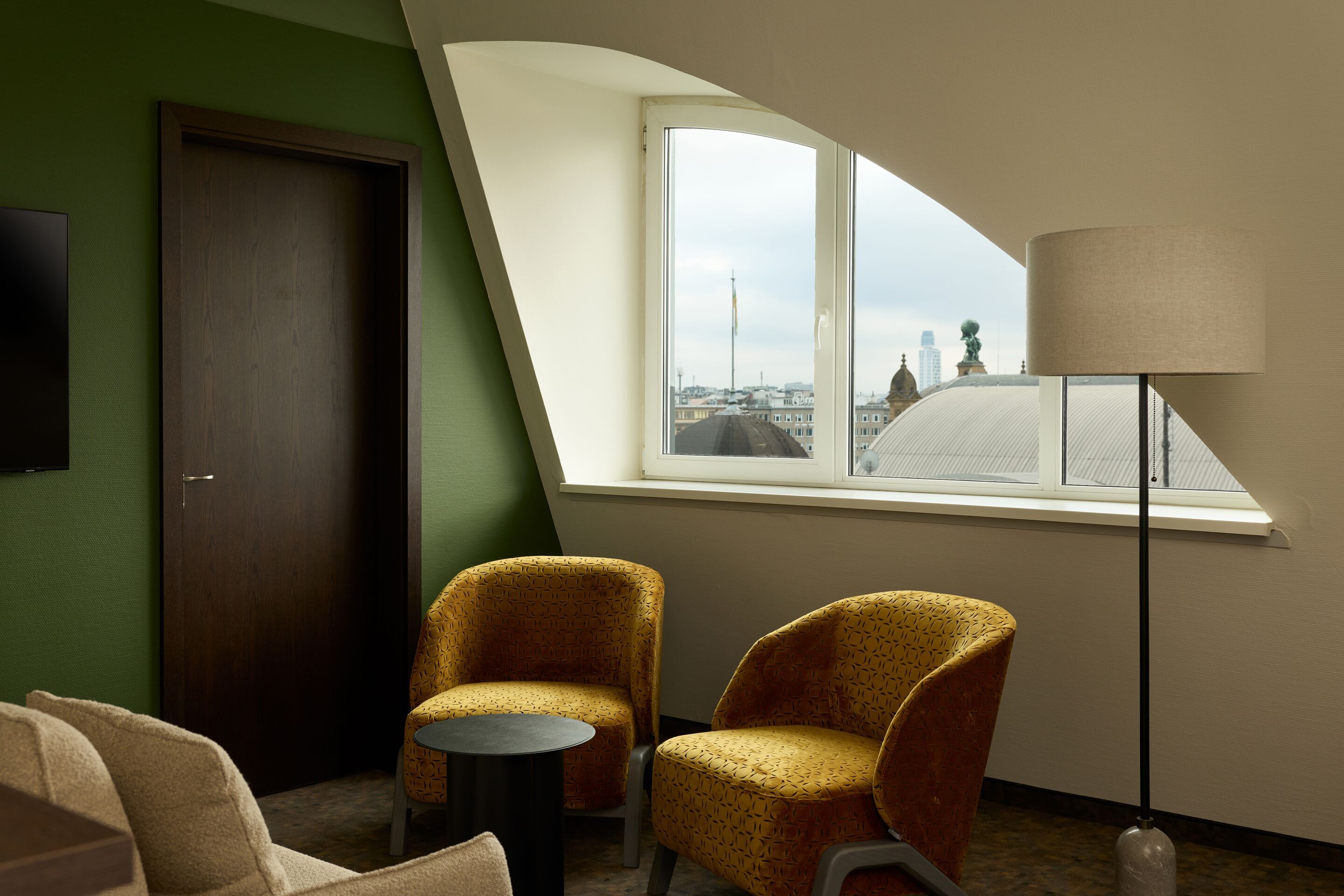 Hotel Frankfurt Metropolitan The Cloud One Zimmer mit gemütlicher Sitzecke mit zwei senfgelben Sesseln, kleinem Tisch und Stehlampe vor großen Fenstern mit Blick auf Stadtdächer, in einem lichtdurchfluteten Hotelzimmer mit grün gestrichener Wand.