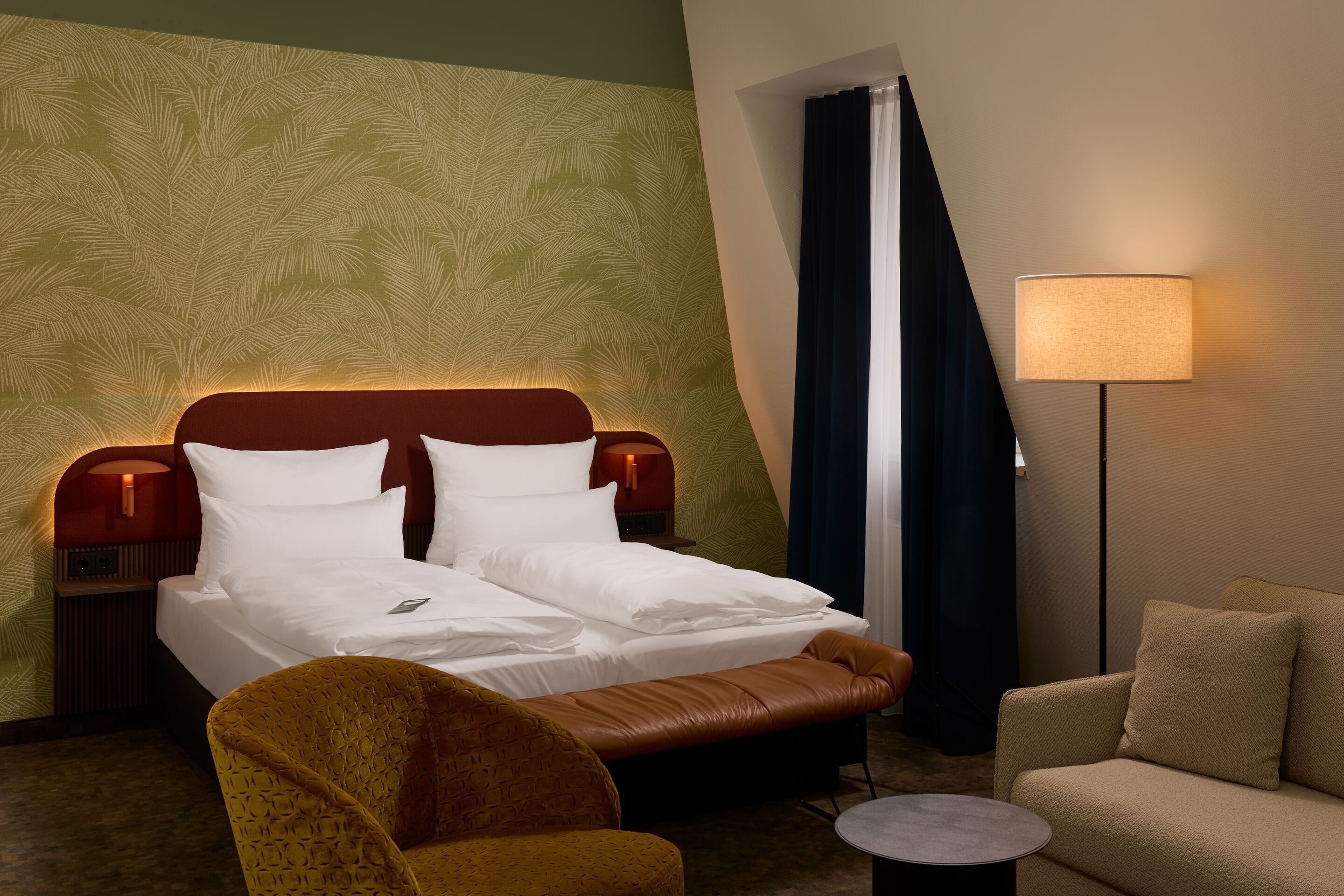 Gemütlich eingerichtetes Hotelzimmer mit Doppelbett, warmem indirektem Licht und gemütlichen Sesseln, neben großem Fenster mit Vorhängen, Atmosphäre lädt zum Entspannen und Wohlfühlen ein.