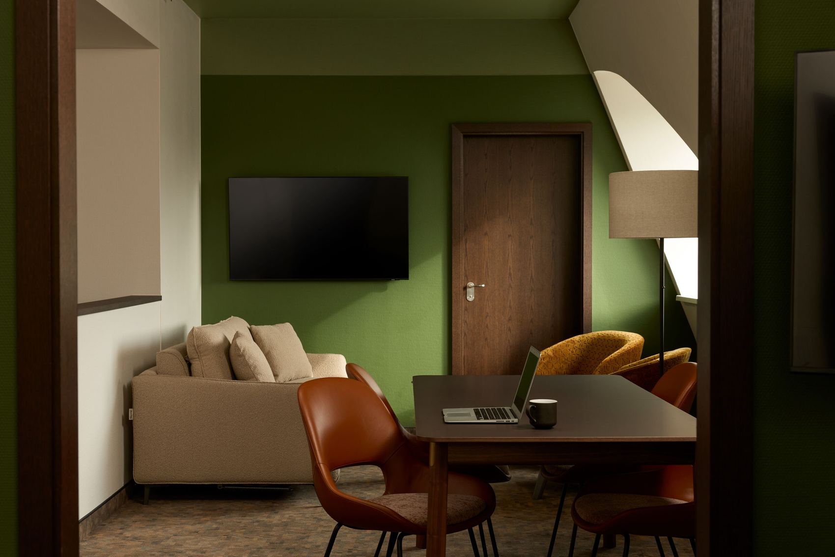 Modern eingerichtetes Hotelzimmer mit grün gestrichener Wand, gemütlichem Sofa, Wandfernseher, Arbeitsplatz mit Laptop und Kaffeetasse sowie komfortablen Stühlen, die eine ruhige und funktionale Atmosphäre für Entspannung und Arbeit schaffen.