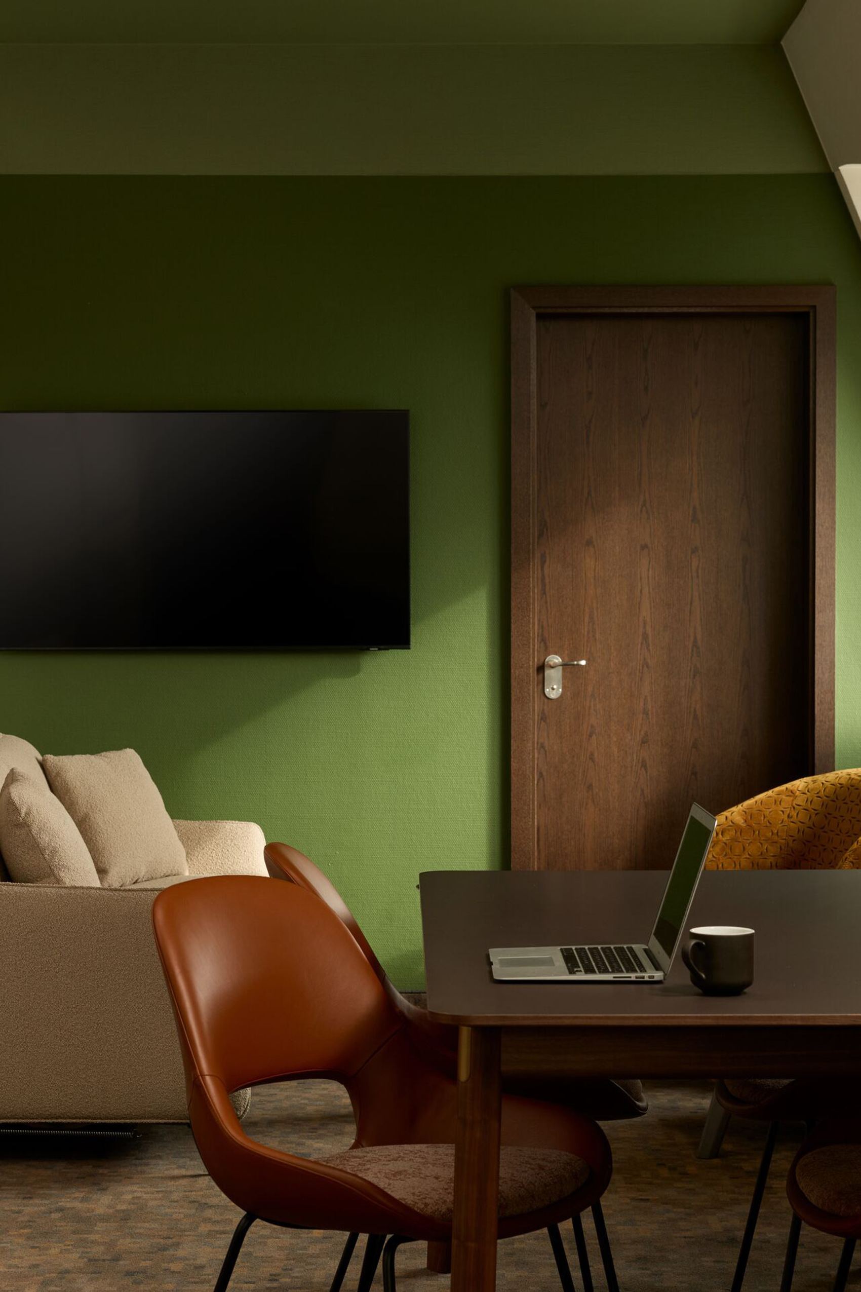 Hotel Frankfurt Metropolitan The Cloud One Zimmer mit grün gestrichener Wand, beigem Sofa, Flachbildfernseher, Holztisch mit Laptop und Kaffeetasse sowie mehreren Stühlen und Lampe.