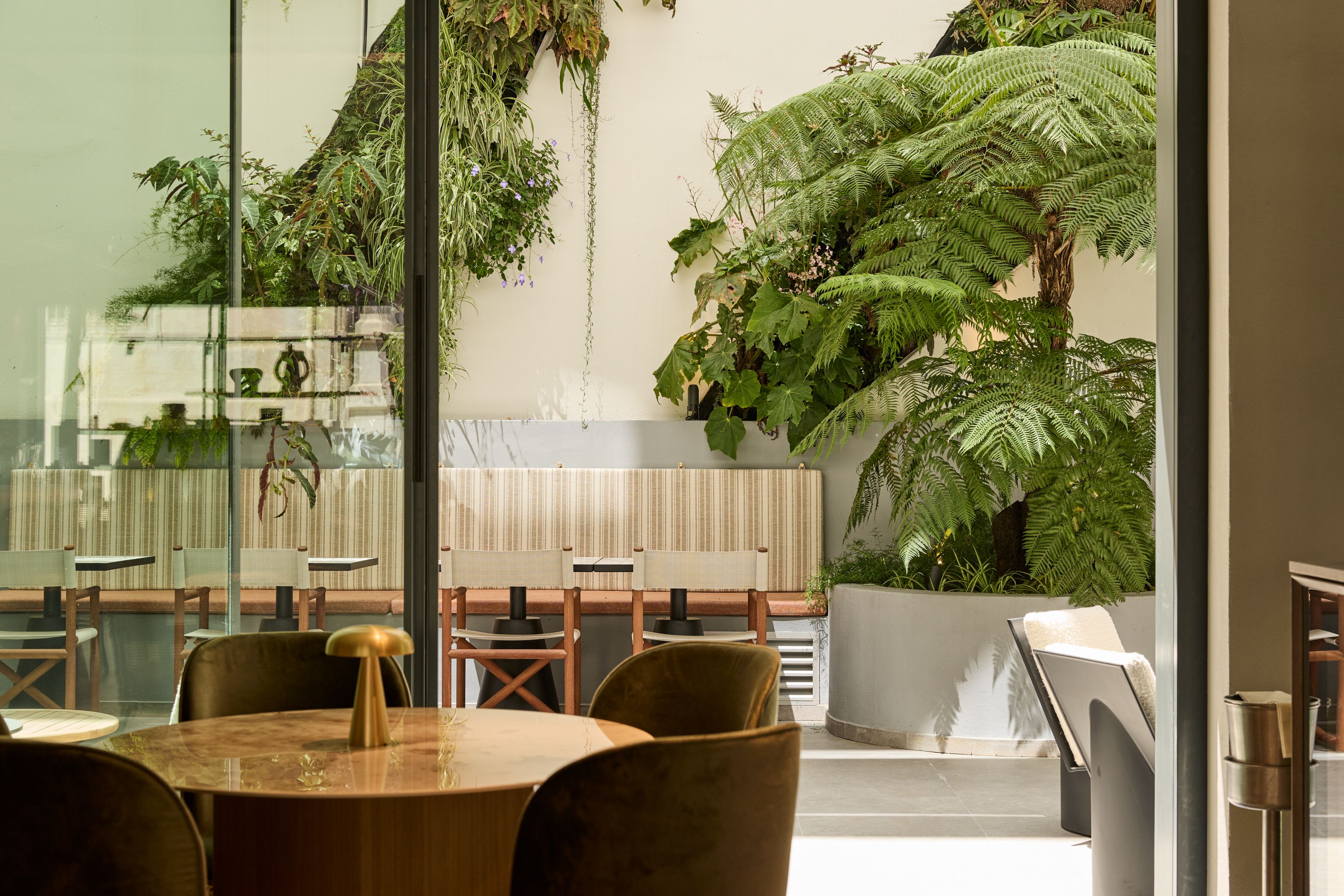 Hotel Lissabon The Cloud One Lounge-Bereich mit runden Tischen, bequemen Stühlen und großer Fensterfront, die auf eine grüne, pflanzenreiche Terrasse hinausblickt.