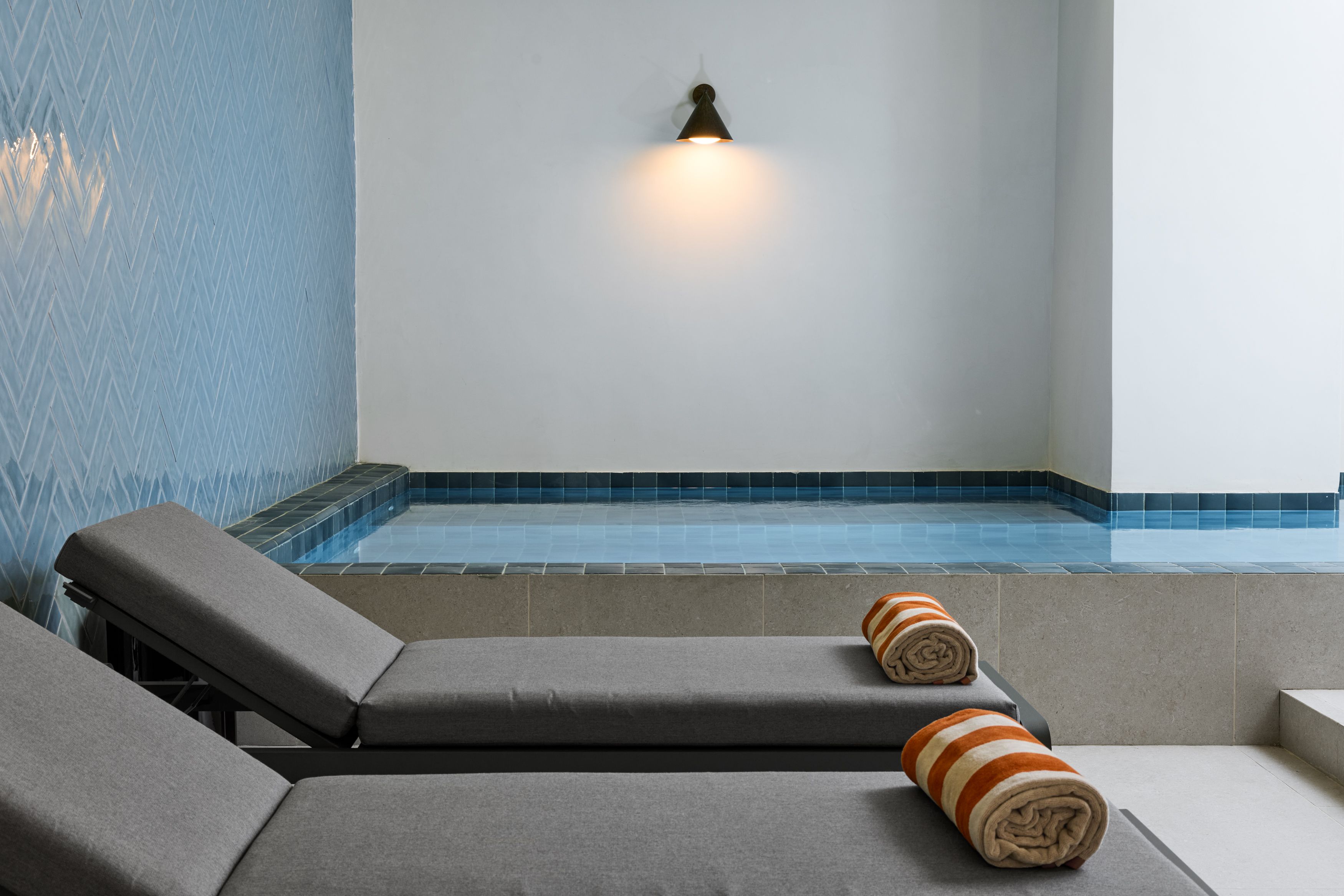 Designhotel Lissabon The Cloud One Pool mit zwei bequemen grauen Liegen im Vordergrund, jeweils mit einem gerollten Handtuch. Im Hintergrund ein mit Wasser gefülltes Becken und eine wandmontierte Lichtquelle.