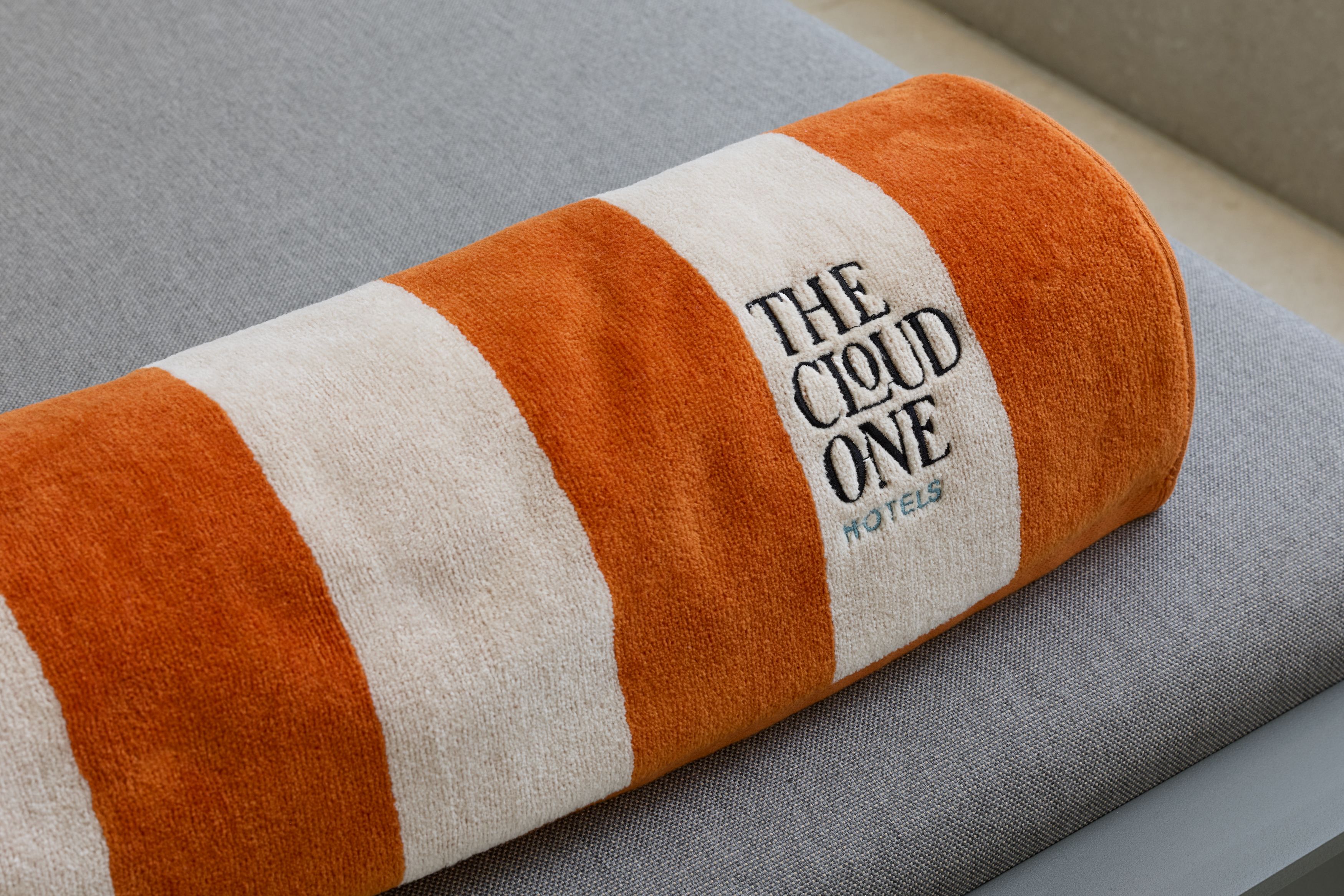 Hotel Lissabon The Cloud One Handtuch mit orange-weißen Streifen und dem Schriftzug „THE CLOUD ONE HOTELS“ liegt auf grauem Polster.