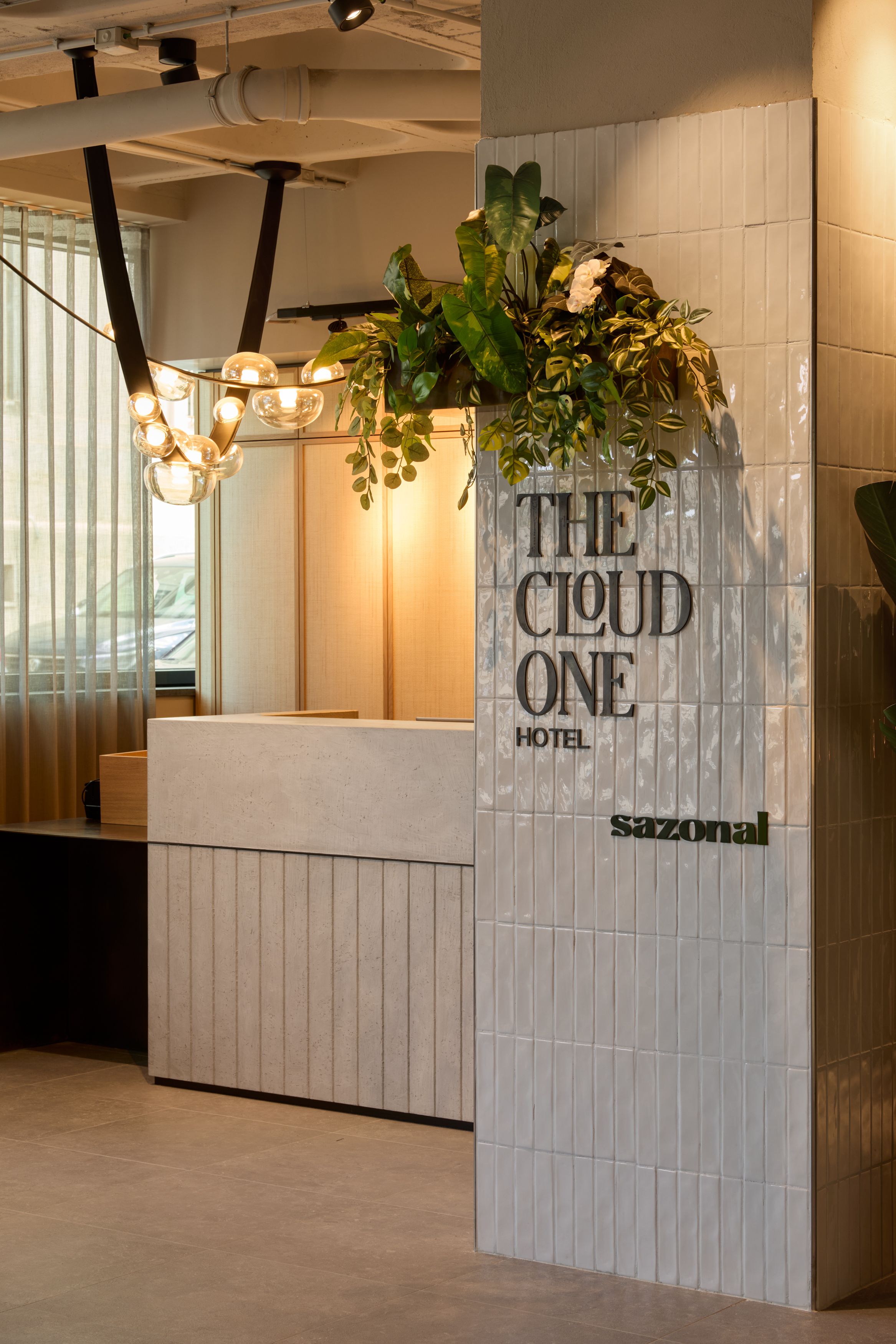 Hotel Lissabon The Cloud One Empfangsbereich mit Rezeption, dekorativen Pflanzen und stilvoller Wandbeschriftung „THE CLOUD ONE HOTEL" und „sazonal“. 