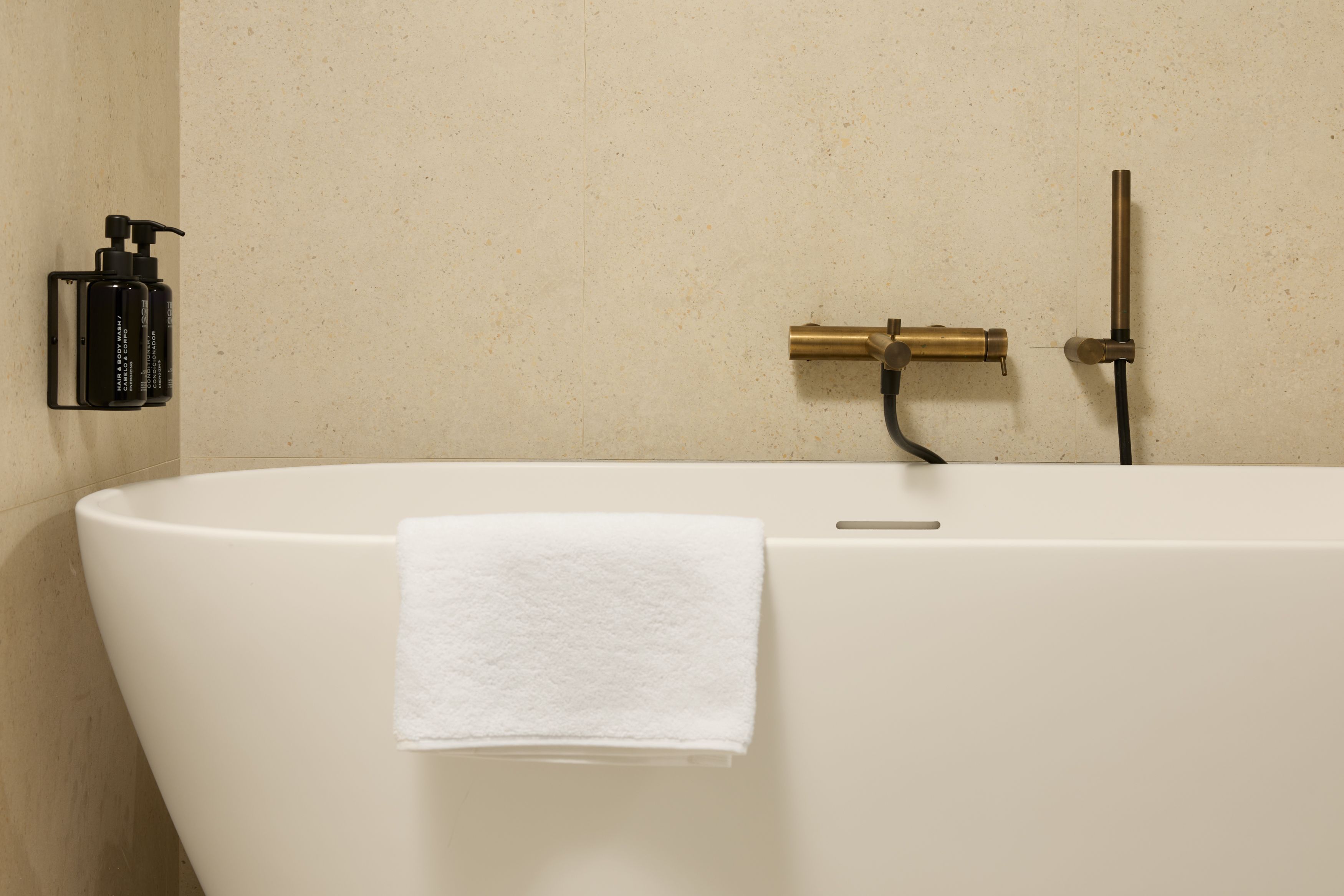 Hotel Lissabon The Cloud One freistehende Badewanne mit weißem Handtuch, minimalistischer Wandarmatur in Messing und Shampoo- sowie Duschgelspender an einer hellen Wand.