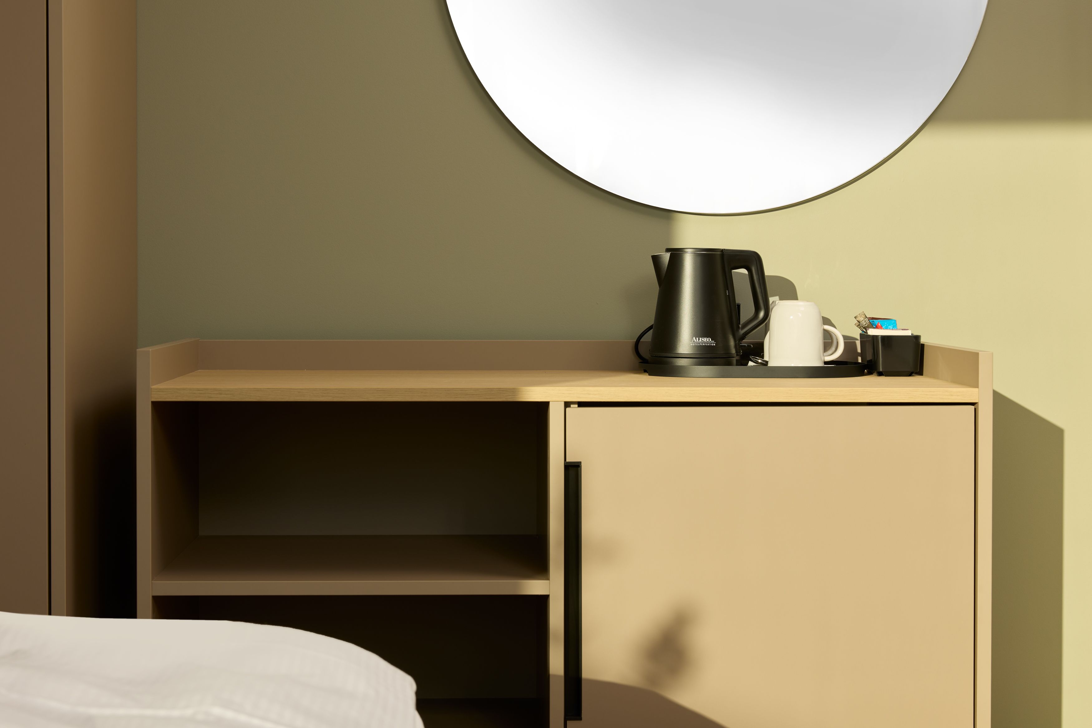 Hotel Lissabon The Cloud One Hotelzimmer mit schlichtem Holzmöbel, elektrischem Wasserkocher, Tasse und Kaffee- oder Teebeuteln auf dem Board, dazu ein großer runder Spiegel an der Wand und weiches Tageslicht.