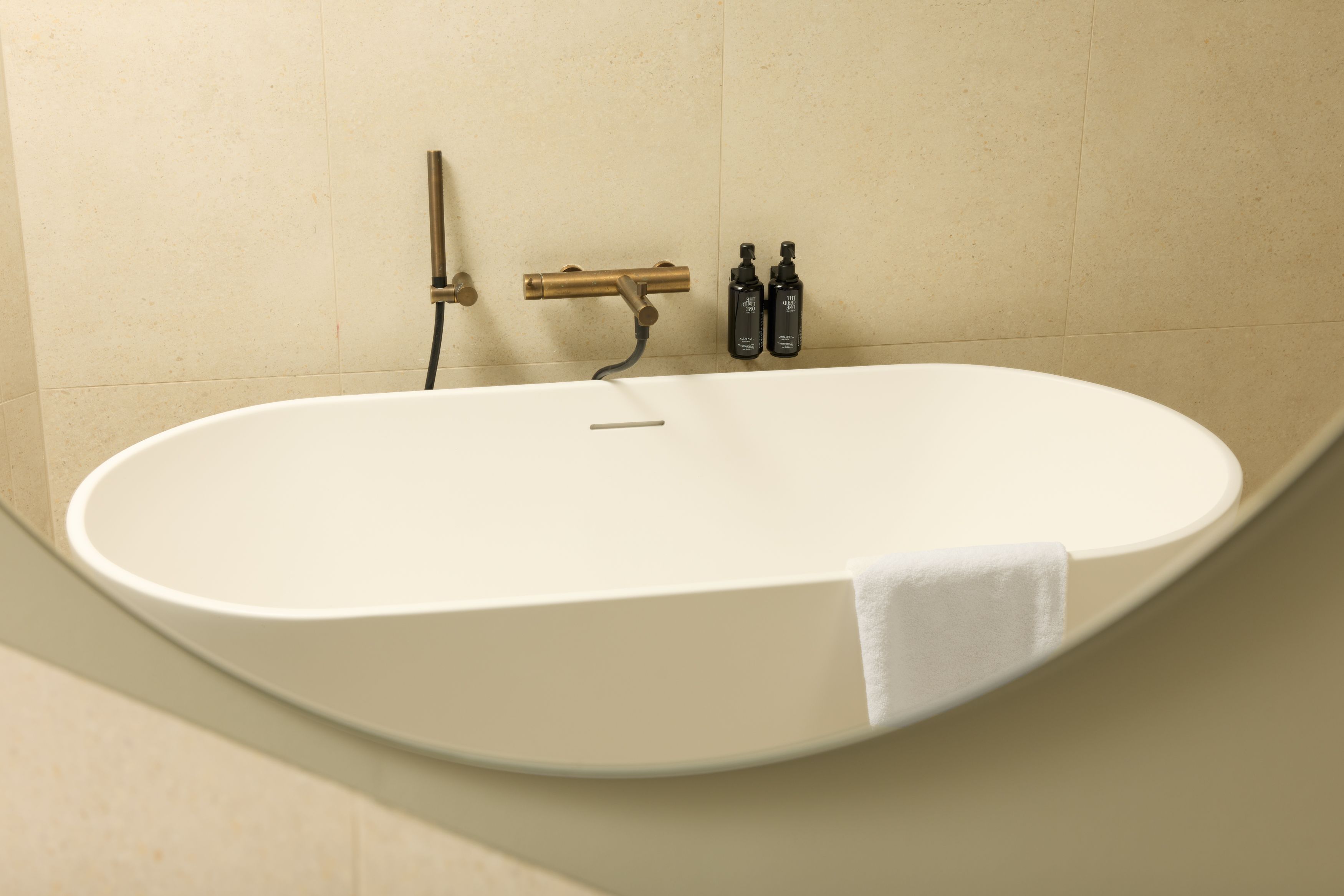 Hotel Lissabon The Cloud One freistehende Badewanne in weiß, modernem Messing-Wasserhahn und weißem Handtuch sowie zwei schwarzen Shampooflaschen, umgeben von warmen Sandsteinfliesen.