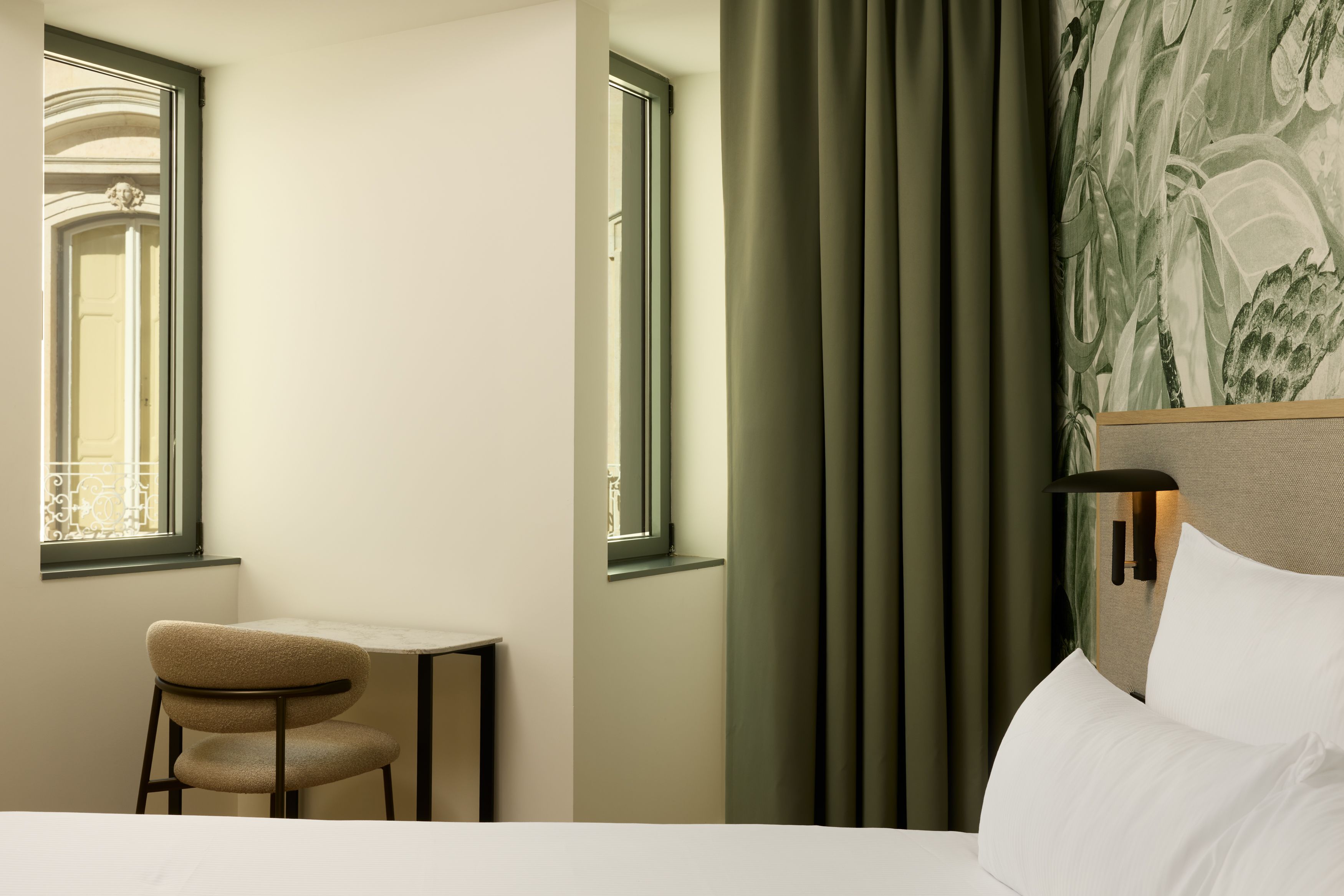 Hotel Lissabon The Cloud One lichtdurchflutetes Zimmer mit großem Bett, grünem Pflanzenmuster an der Wand, dunkelgrünen Vorhängen und einem kleinen Schreibtisch mit Stuhl vor zwei Fenstern.