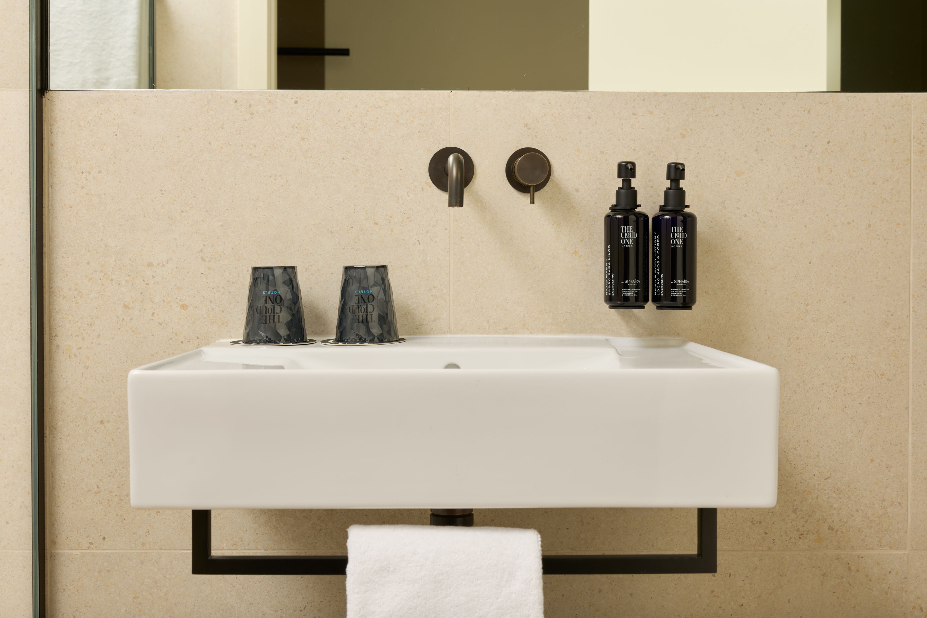 Designhotel Lissabon The Cloud One Waschtisch mit schwarzem Handtuchhalter unter dem Waschbecken, zwei dunklen Trinkgläsern und befestigten Spendern für Seife und Lotion, kombiniert mit wandmontierten Armaturen in einem hellen Badezimmer.
