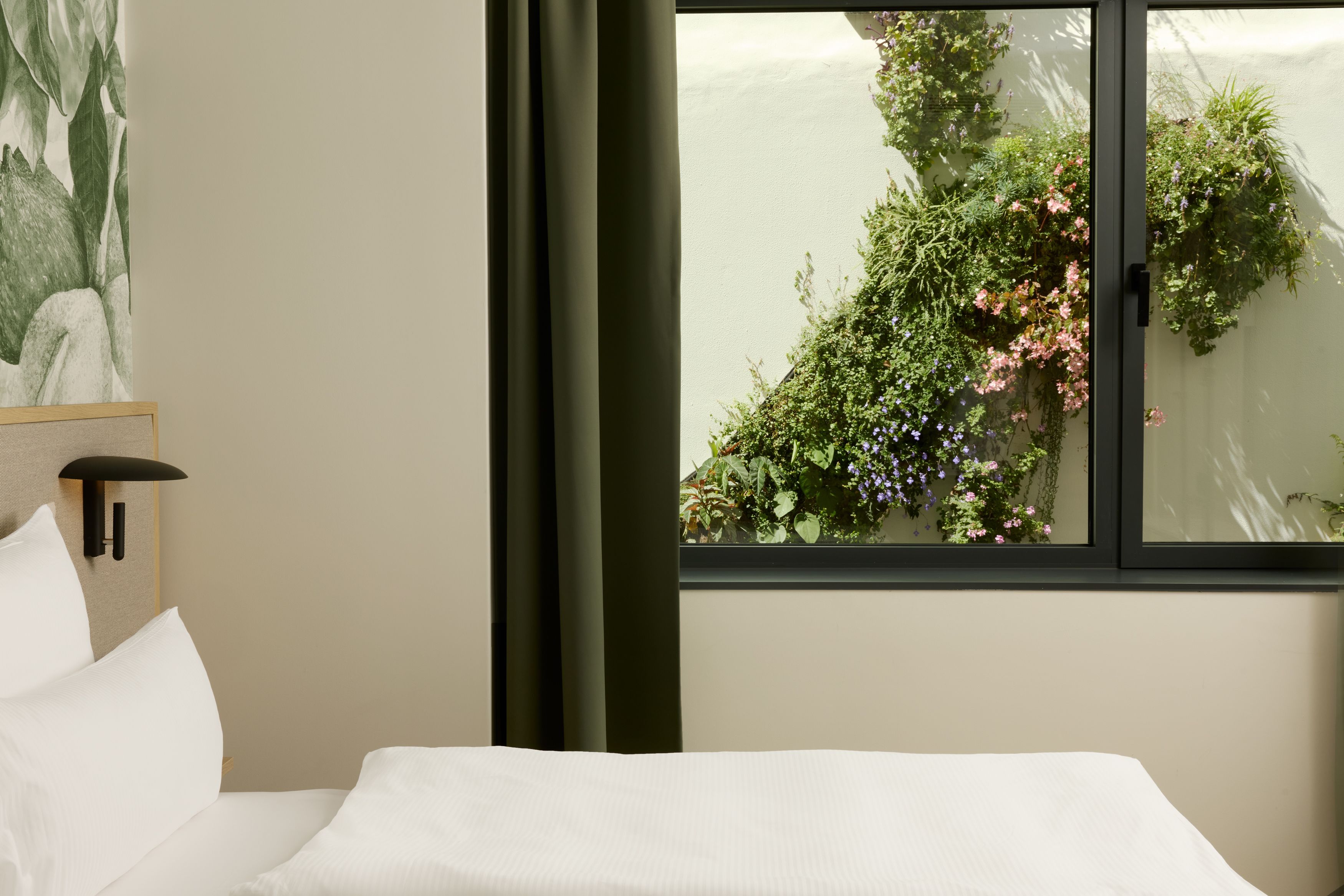 Hotel Lissabon The Cloud One hell und modern eingerichtetes Hotelzimmer mit großem Fenster, das den Blick auf eine blühende vertikale Gartenwand im Innenhof bietet, und einem gemütlichen, weißen Bett.