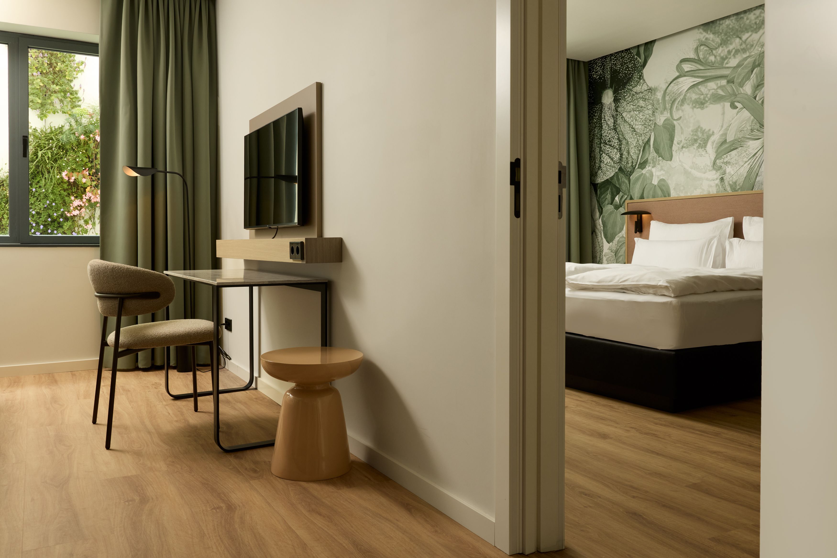 Hotel Lissabon The Cloud One Hotelzimmer mit Holzfußboden, Spiegel, Arbeitsbereich, Flachbildfernseher und angrenzendem Schlafzimmer mit gemütlichem Bett und natürlicher Wandgestaltung.