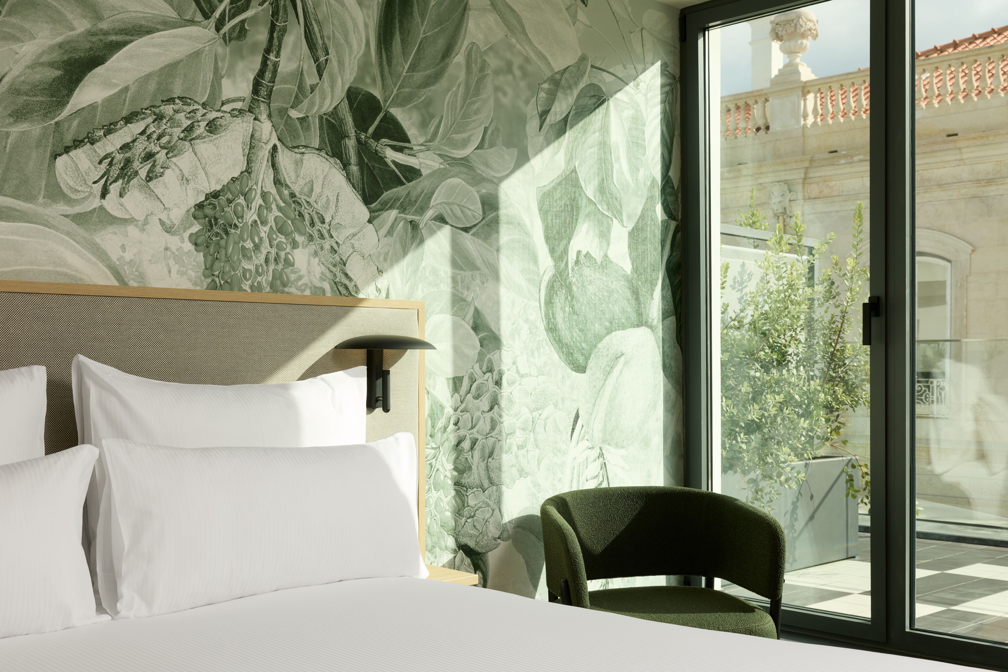 Hotel Lissabon The Cloud One lichtdurchflutetes Hotelzimmer mit großem, bequemem Bett, floral gestalteter Wand, grünem Sessel und bodentiefem Fenster mit Blick auf den sonnigen Balkon. 