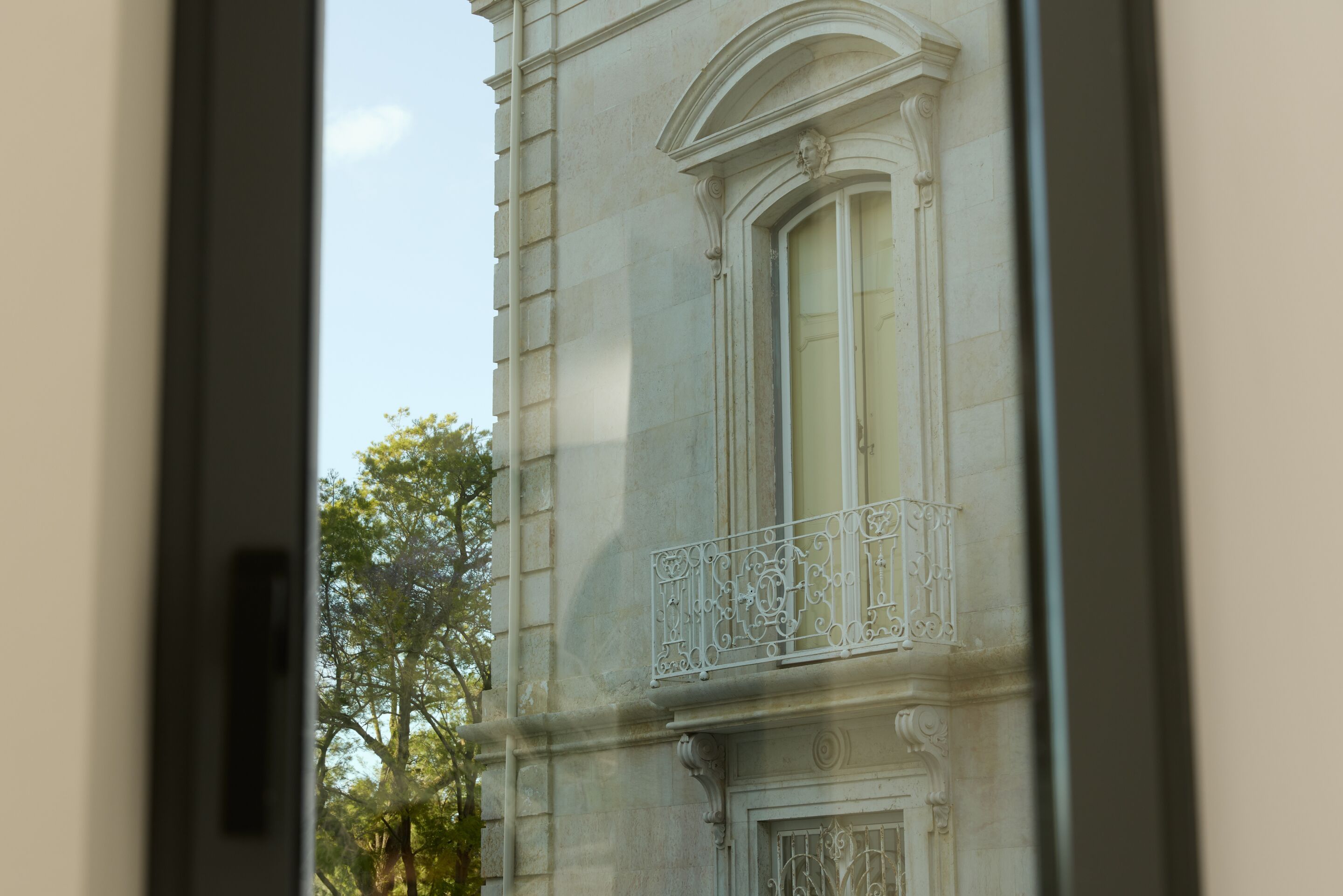 The Cloud One Lisbon Blick durch ein geöffnetes Fenster auf die Fassade eines historischen Gebäudes mit verzierten Balkonbrüstungen und umliegendem Grün im Hintergrund bei Tageslicht.