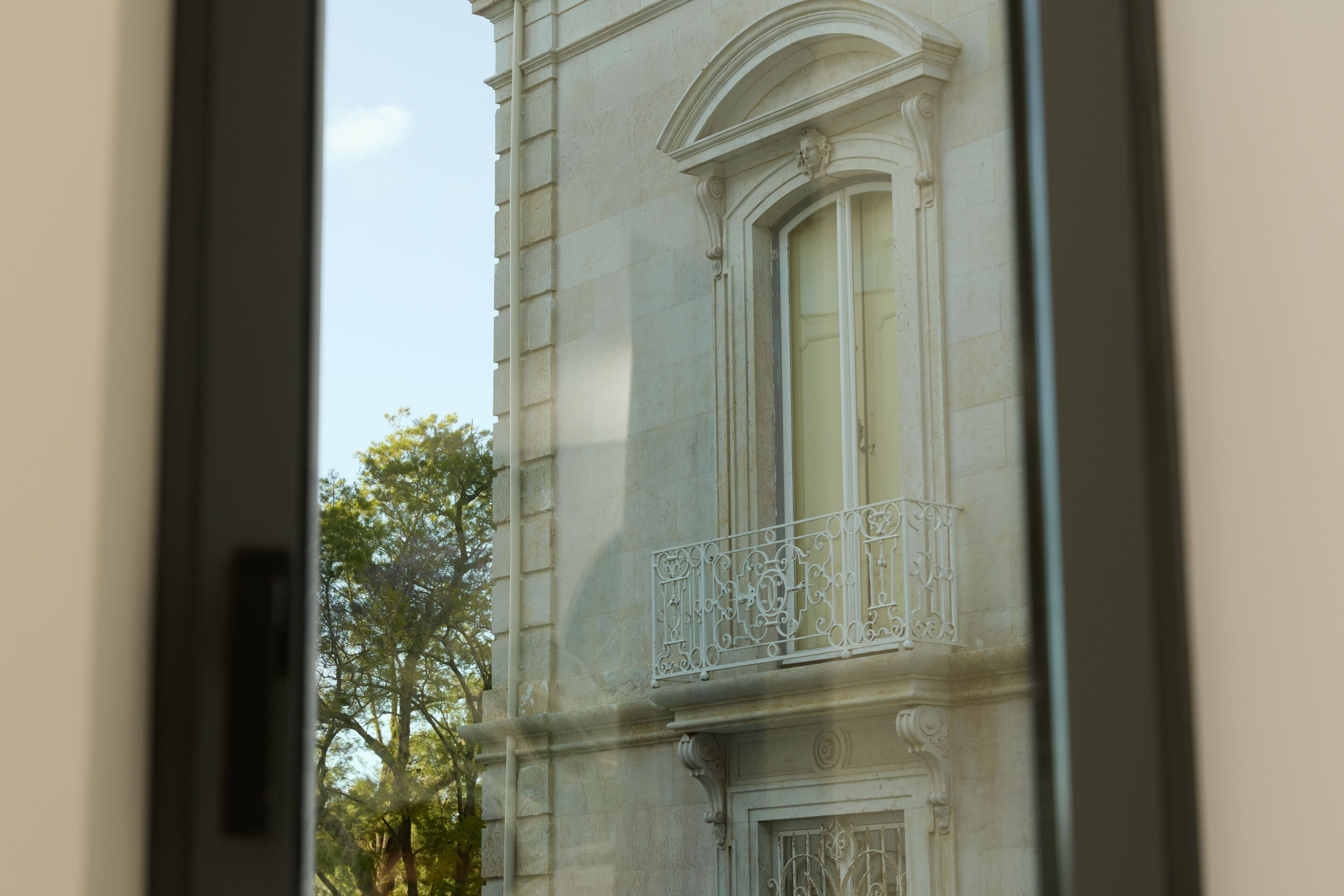 The Cloud One Lisbon Blick durch ein geöffnetes Fenster auf die Fassade eines historischen Gebäudes mit verzierten Balkonbrüstungen und umliegendem Grün im Hintergrund bei Tageslicht. The Cloud One Lisbon Blick durch ein geöffnetes Fenster auf die Fassade eines historischen Gebäudes mit verzierten Balkonbrüstungen und umliegendem Grün im Hintergrund bei Tageslicht.