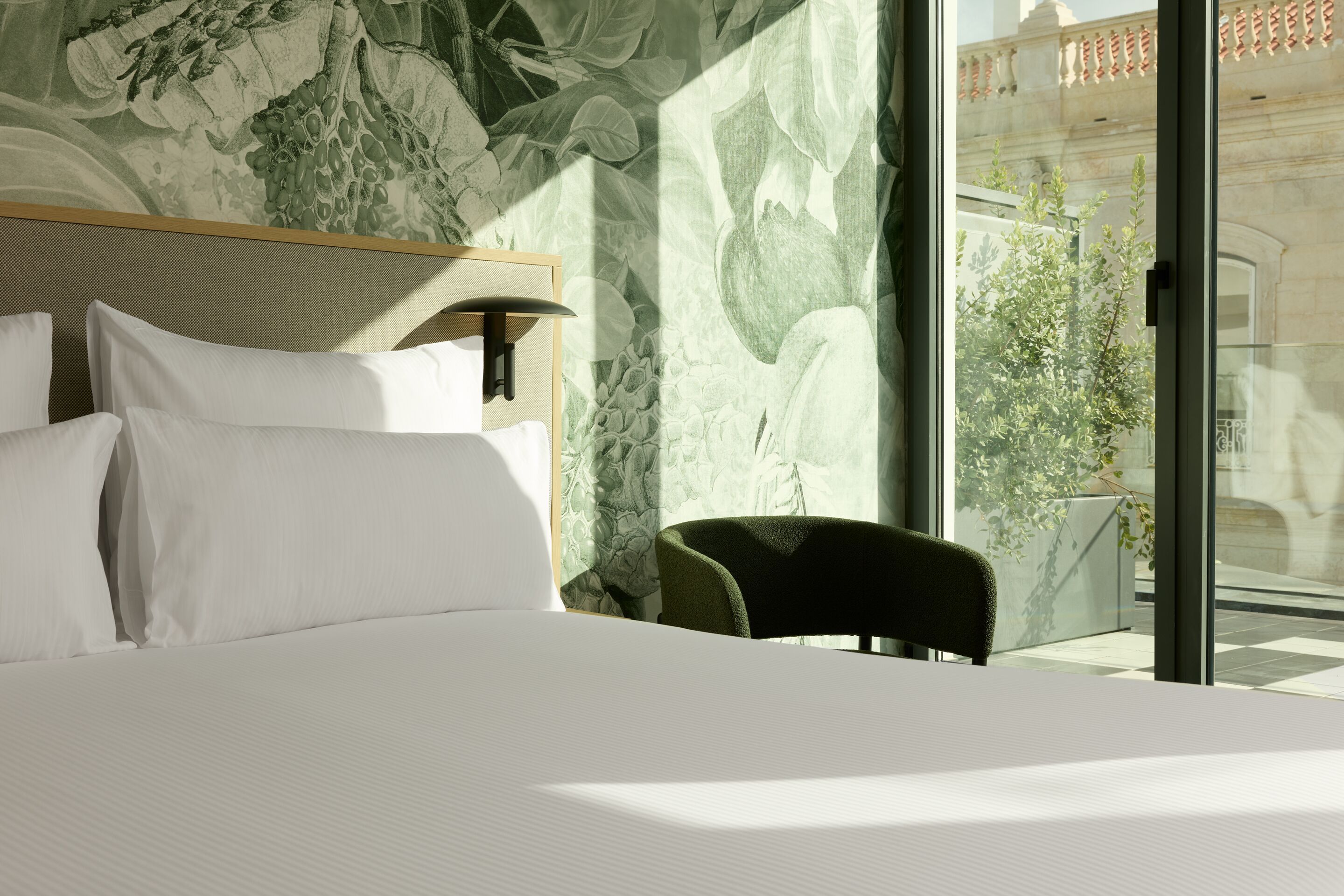 The Cloud One Lisbon Helles, modernes Hotelzimmer mit großem, weiß bezogenem Bett, grünem Akzentstuhl und bodentiefem Fenster, das auf einen Balkon mit Blick ins Grüne öffnet. Eine Wand ist dekorativ mit Pflanzenmotiven gestaltet, die Raum und Atmosphäre natürliche Frische verleihen.