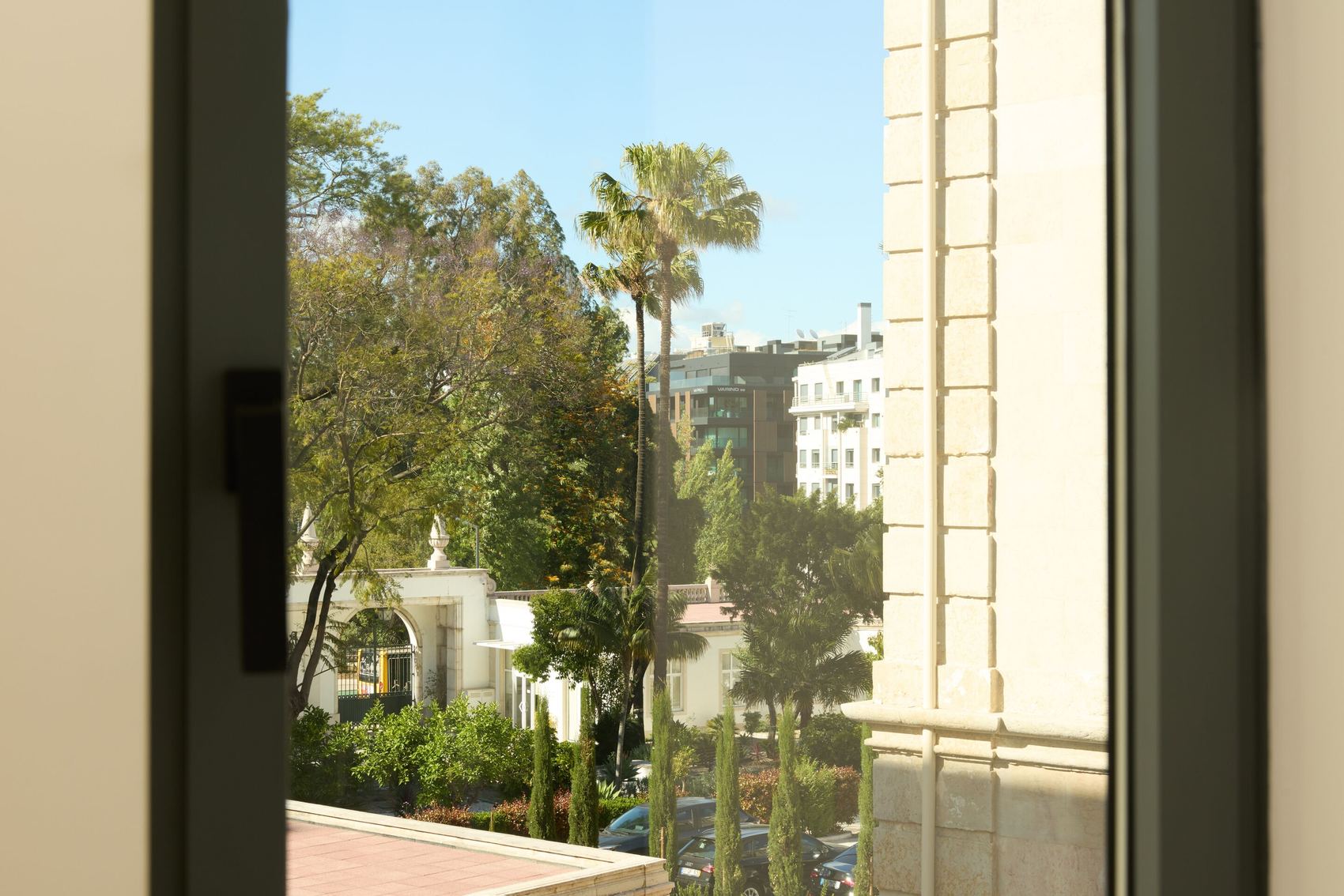 The Cloud One Lisbon Blick aus einem Fenster auf einen hellen Innenhof mit Palmen, grünen Bäumen und Gebäuden im Hintergrund bei klarem Himmel.