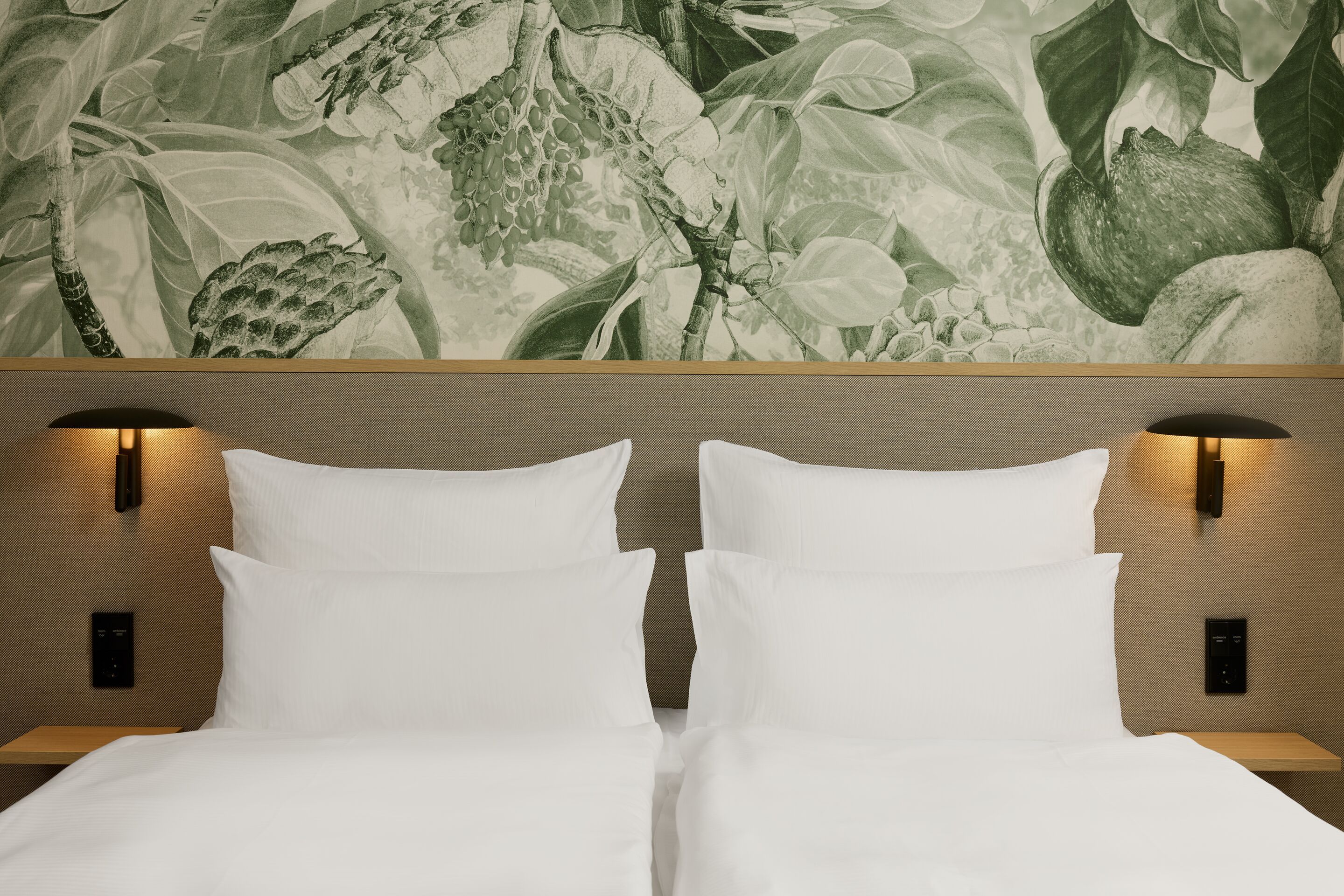 The Cloud One Lisbon Zwei Einzelbetten mit weißen Bettlaken und Kissen, seitlich beleuchtet von modernen Wandlampen, vor einer Wand mit stilisiertem Pflanzen- und Fruchtmuster, die eine ruhige und naturnahe Atmosphäre im Raum schafft.