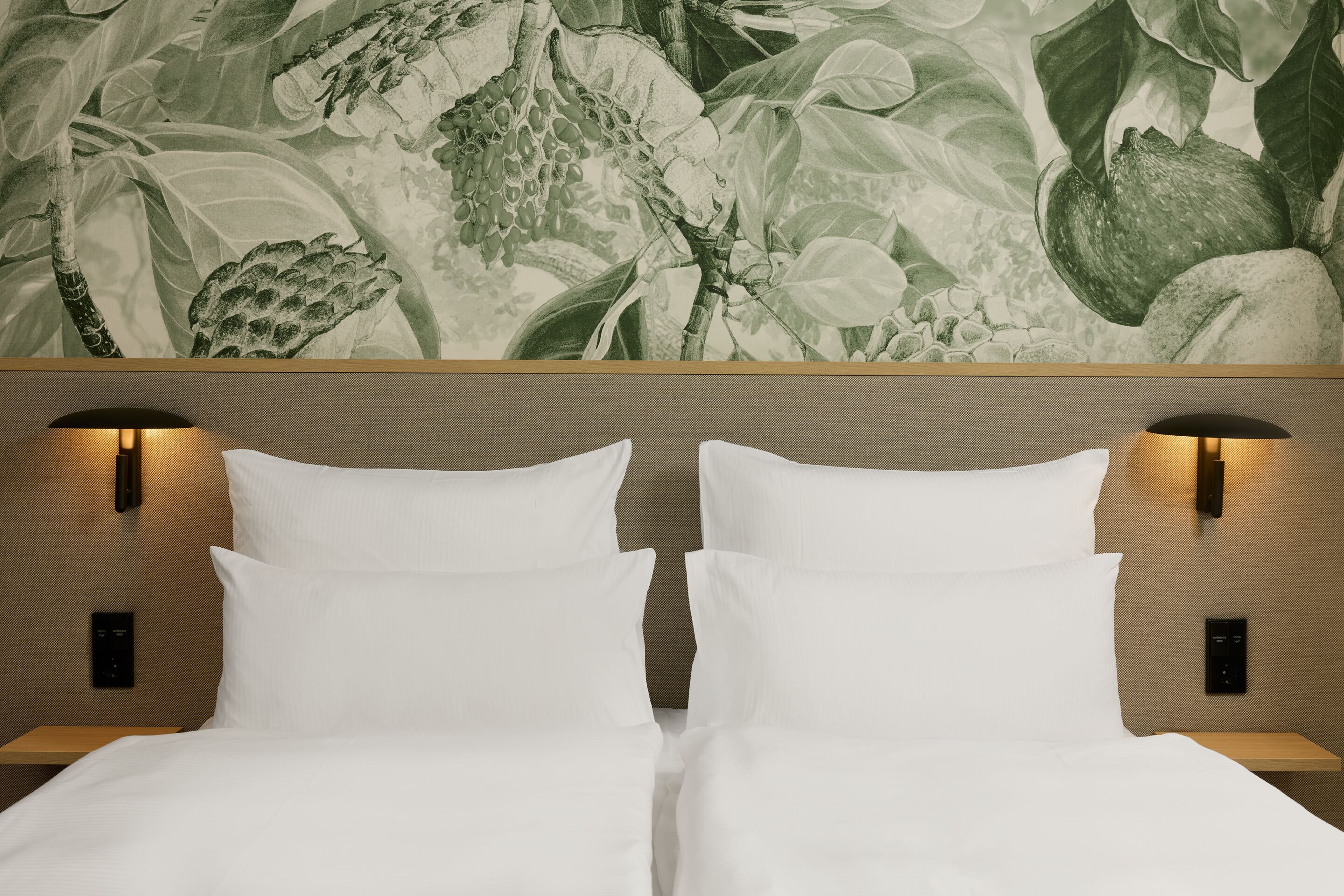 The Cloud One Lisbon Zwei Einzelbetten mit weißen Bettlaken und Kissen, seitlich beleuchtet von modernen Wandlampen, vor einer Wand mit stilisiertem Pflanzen- und Fruchtmuster, die eine ruhige und naturnahe Atmosphäre im Raum schafft. The Cloud One Lisbon Zwei Einzelbetten mit weißen Bettlaken und Kissen, seitlich beleuchtet von modernen Wandlampen, vor einer Wand mit stilisiertem Pflanzen- und Fruchtmuster, die eine ruhige und naturnahe Atmosphäre im Raum schafft.