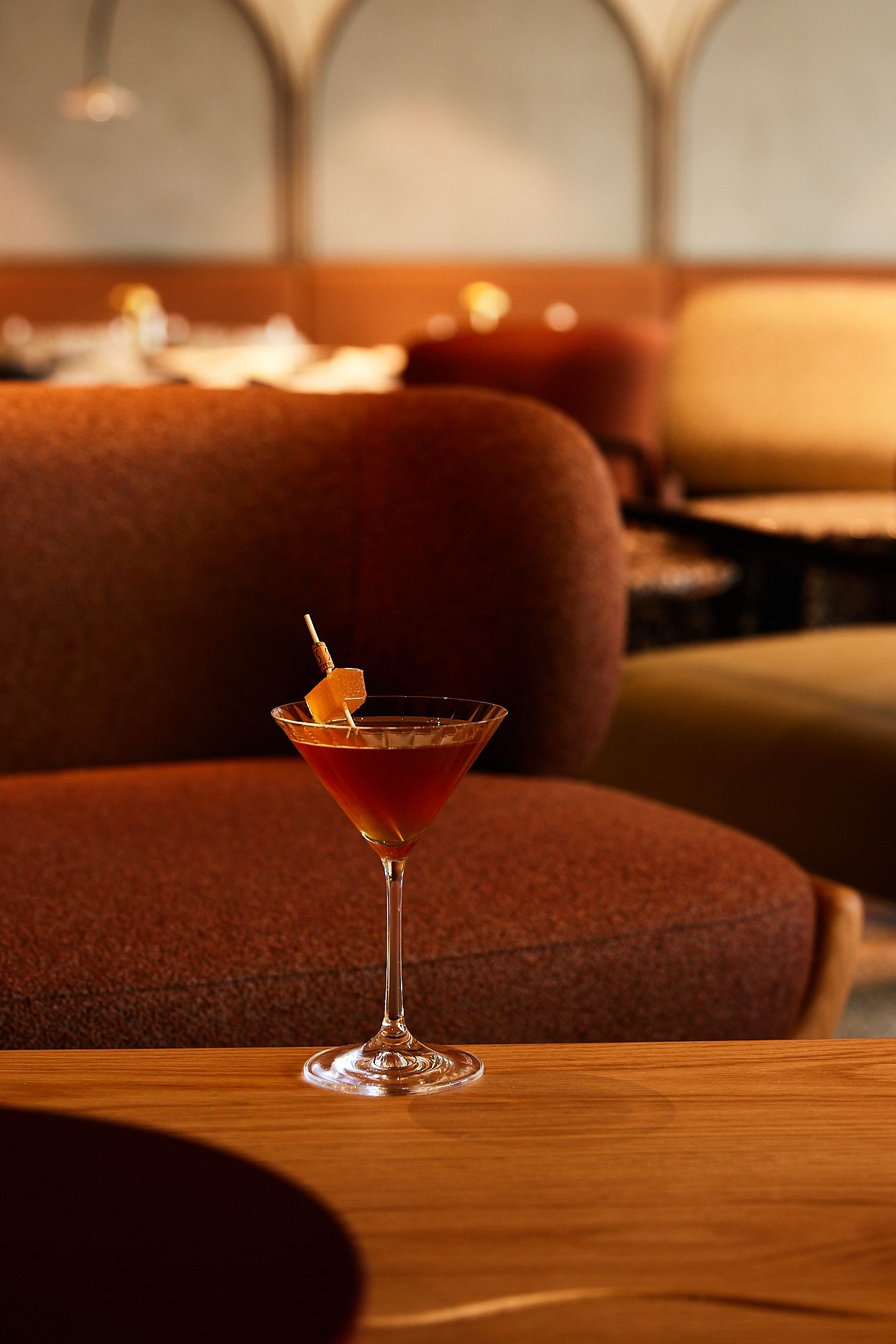 Ein eleganter Cocktail in einem Martiniglas steht auf einem Holztisch vor einer gemütlichen, braunen Sofa- und Sitzgruppe in einer stilvollen Hotelbar-Atmosphäre.