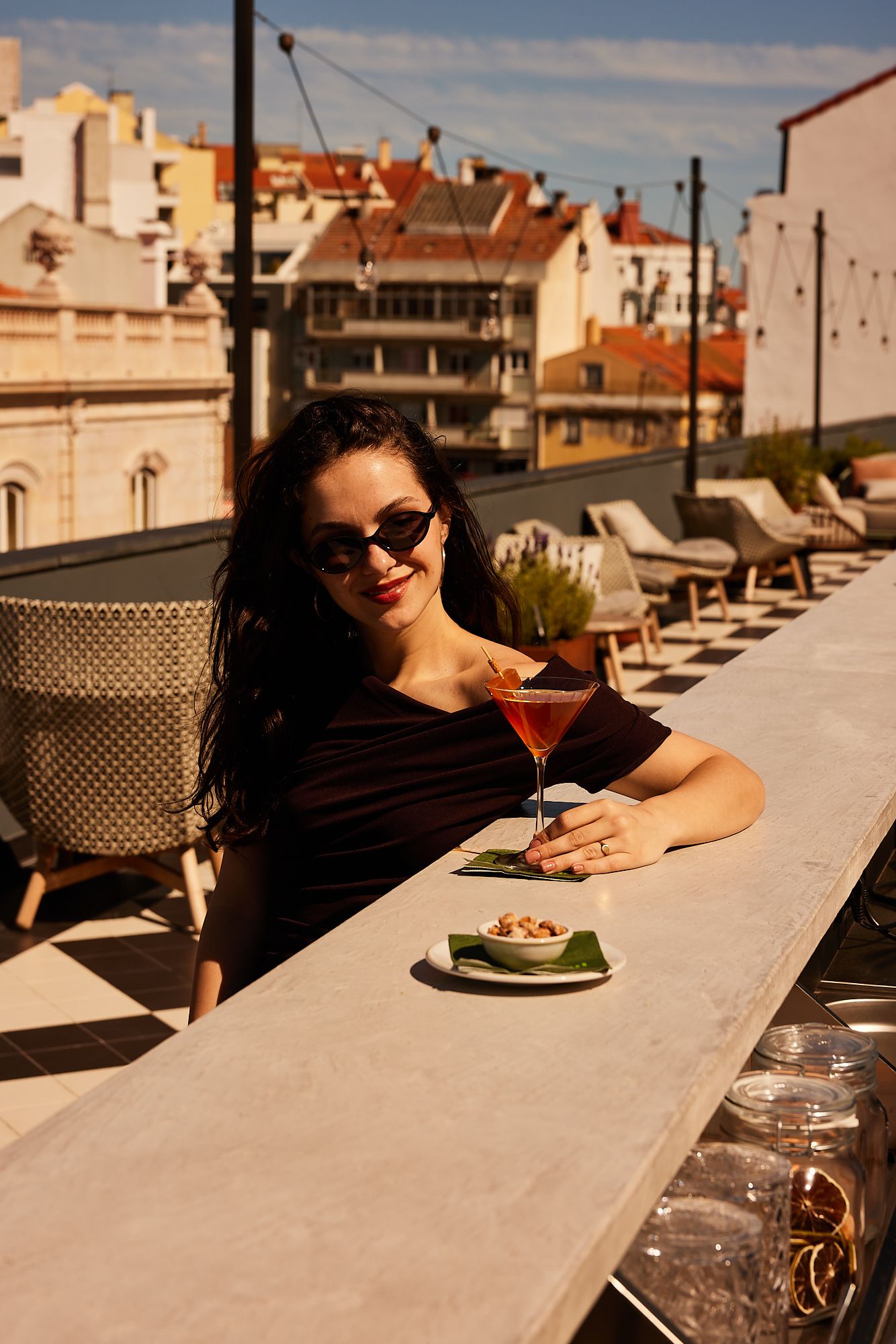 Frau sitzt entspannt auf einer Dachterrasse, genießt bei Sonnenschein einen Cocktail und eine kleine Snackschale mit Blick auf die Stadt.