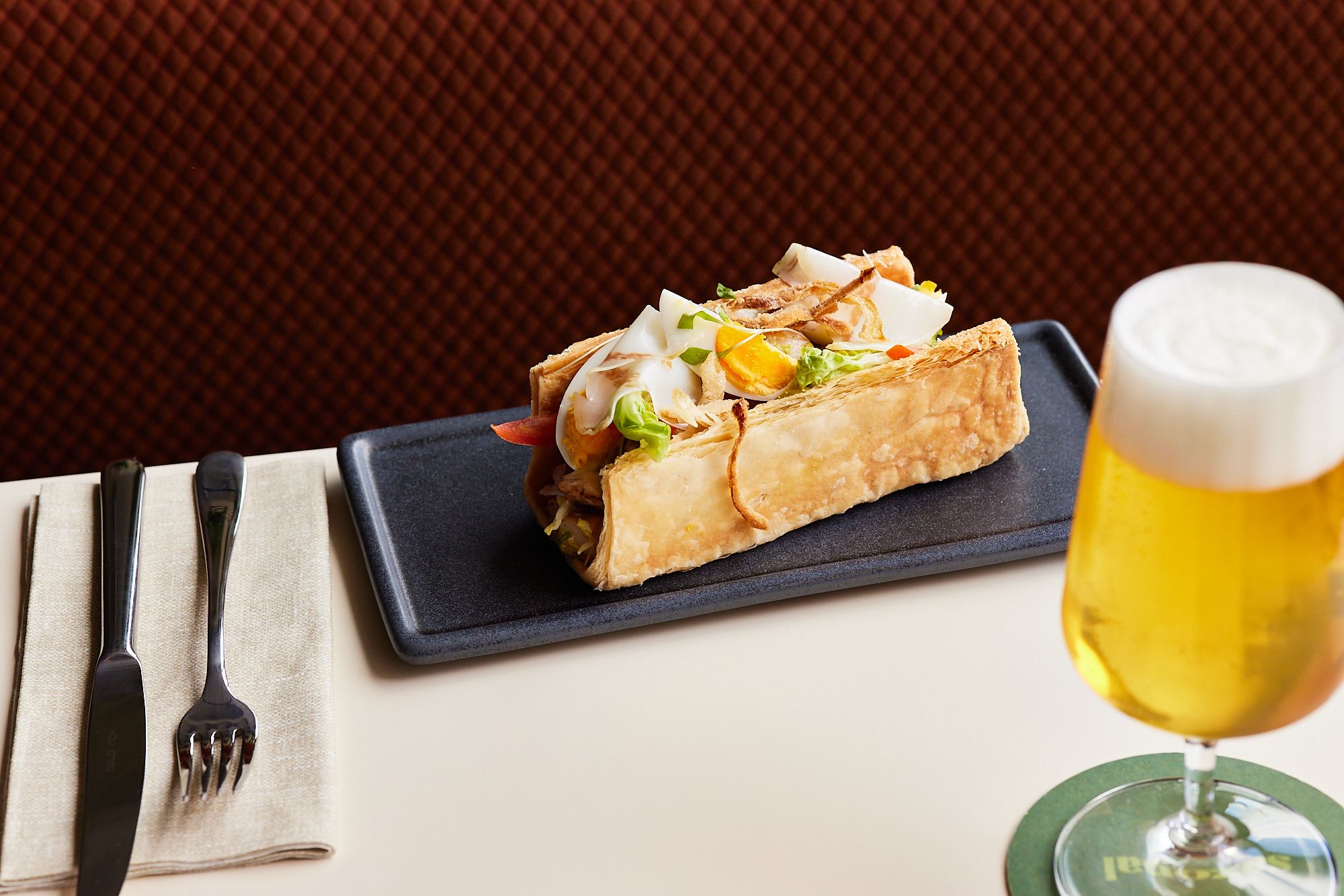Ein belegtes Sandwich mit frischen Zutaten auf einem schwarzen Teller, daneben ein Glas Bier mit Schaum, Besteck und Stoffserviette auf hellem Tisch.