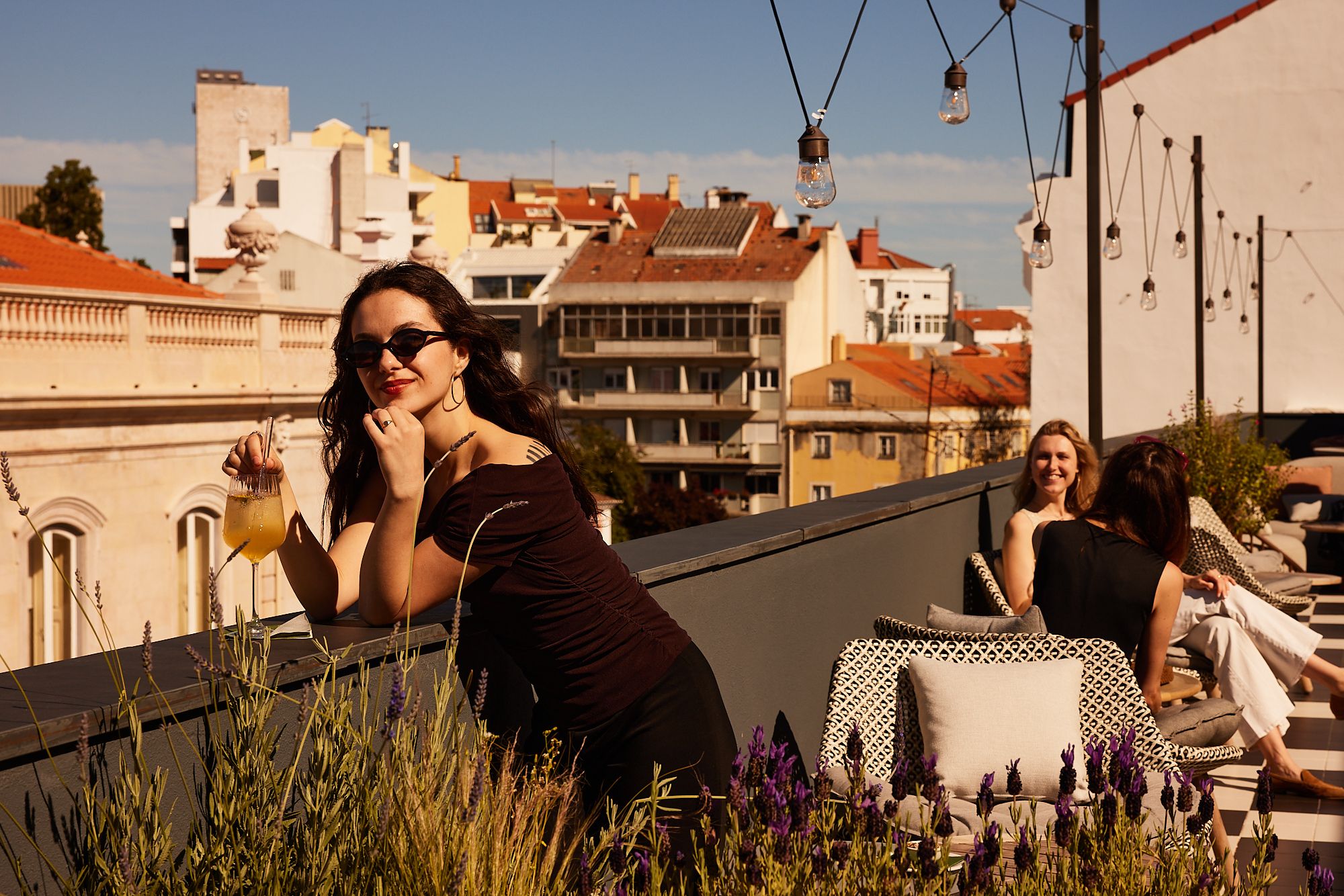 Drei Frauen entspannen auf einer sonnigen Dachterrasse mit Blick auf die Stadt, eine hält ein Getränk, Atmosphäre freundlich und ruhig.