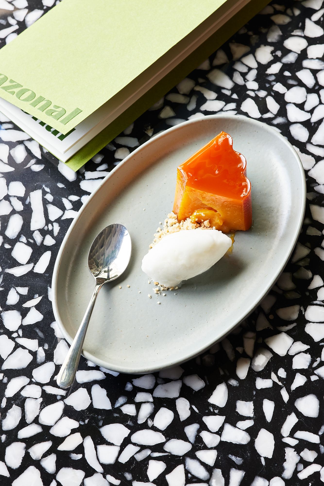 Ein kleines Dessert mit einem Stück Karamellkuchen und einer Kugel Vanilleeis, serviert auf einem ovalen weißen Teller mit einem Löffel daneben, auf einer schwarz-weißen Marmoroberfläche. Daneben liegt ein grünliches Buch.