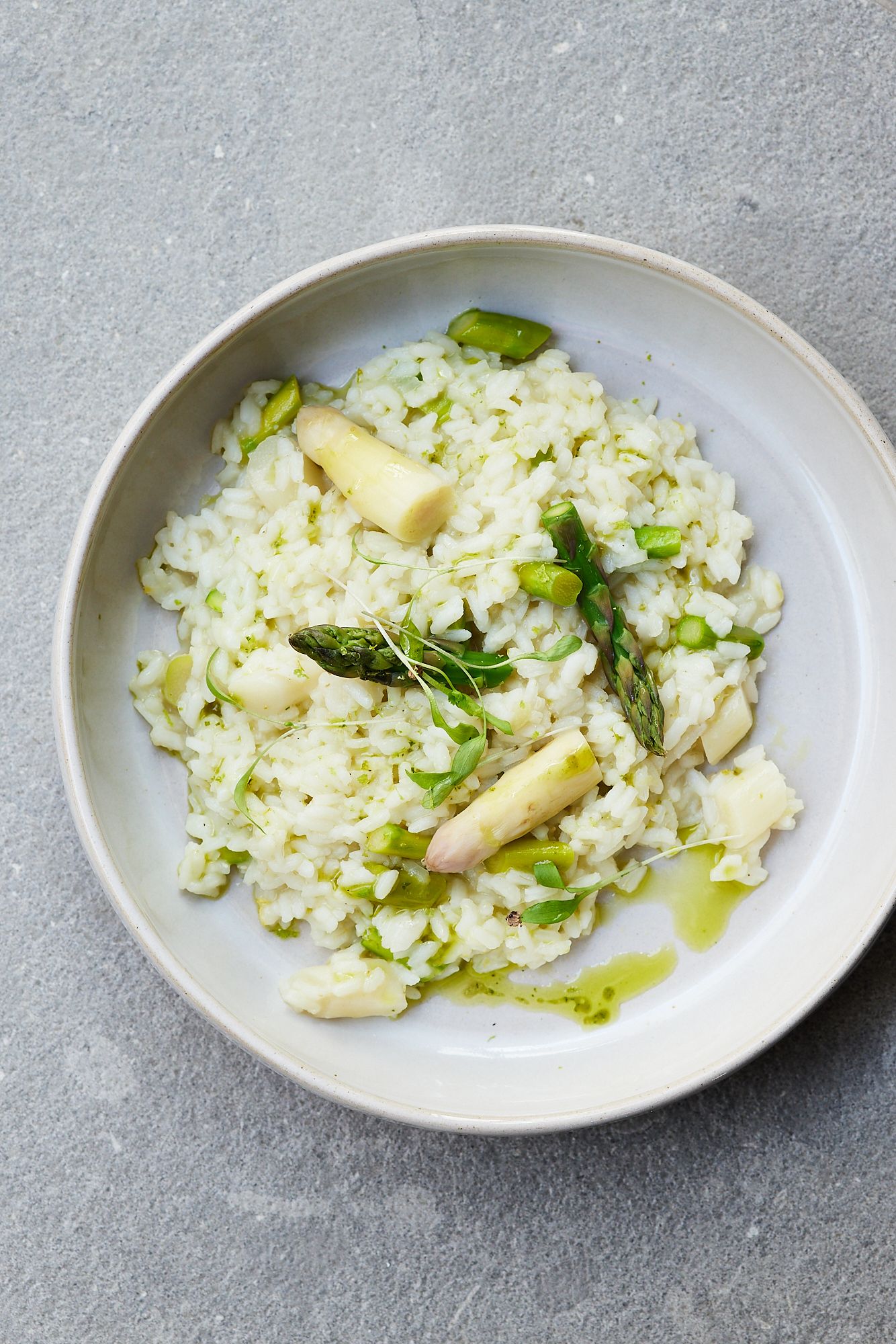 Ein Teller cremiges Risotto mit grünen und weißen Spargelspitzen, garniert mit frischen Kräutern und einem Hauch Olivenöl, serviert auf einem grauen Untergrund.
