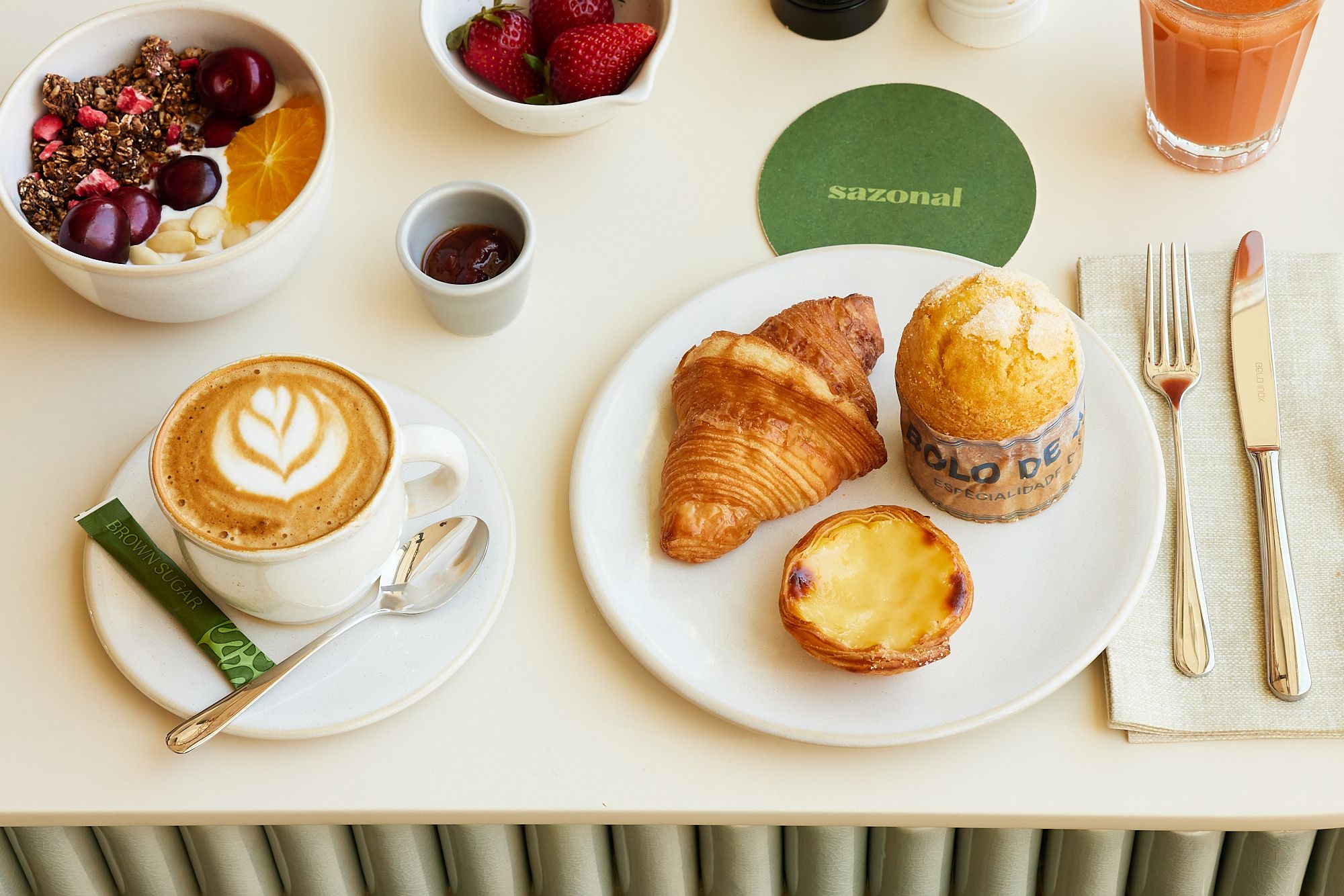Frühstück mit Cappuccino, Croissant, Pudding-Törtchen, Muffin, frischen Erdbeeren, Joghurt mit Granola und Früchten sowie einem Glas Saft, stilvoll auf hellem Tisch angerichtet.