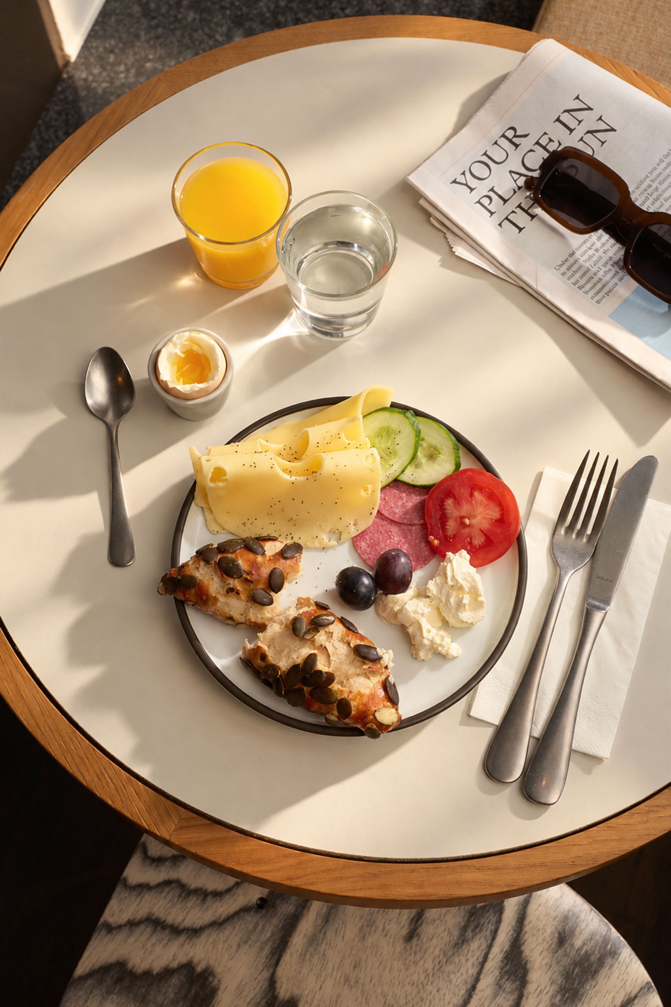 Ein Frühstücksteller mit Vollkornbrötchen, Käse, Gurkenscheiben, Tomate, Trauben und Frischkäse, dazu ein Glas Orangensaft, ein Glas Wasser und ein gekochtes Ei. Neben dem Teller liegen Besteck, ein Löffel, eine Zeitung und eine Sonnenbrille auf einem runden Tisch.
