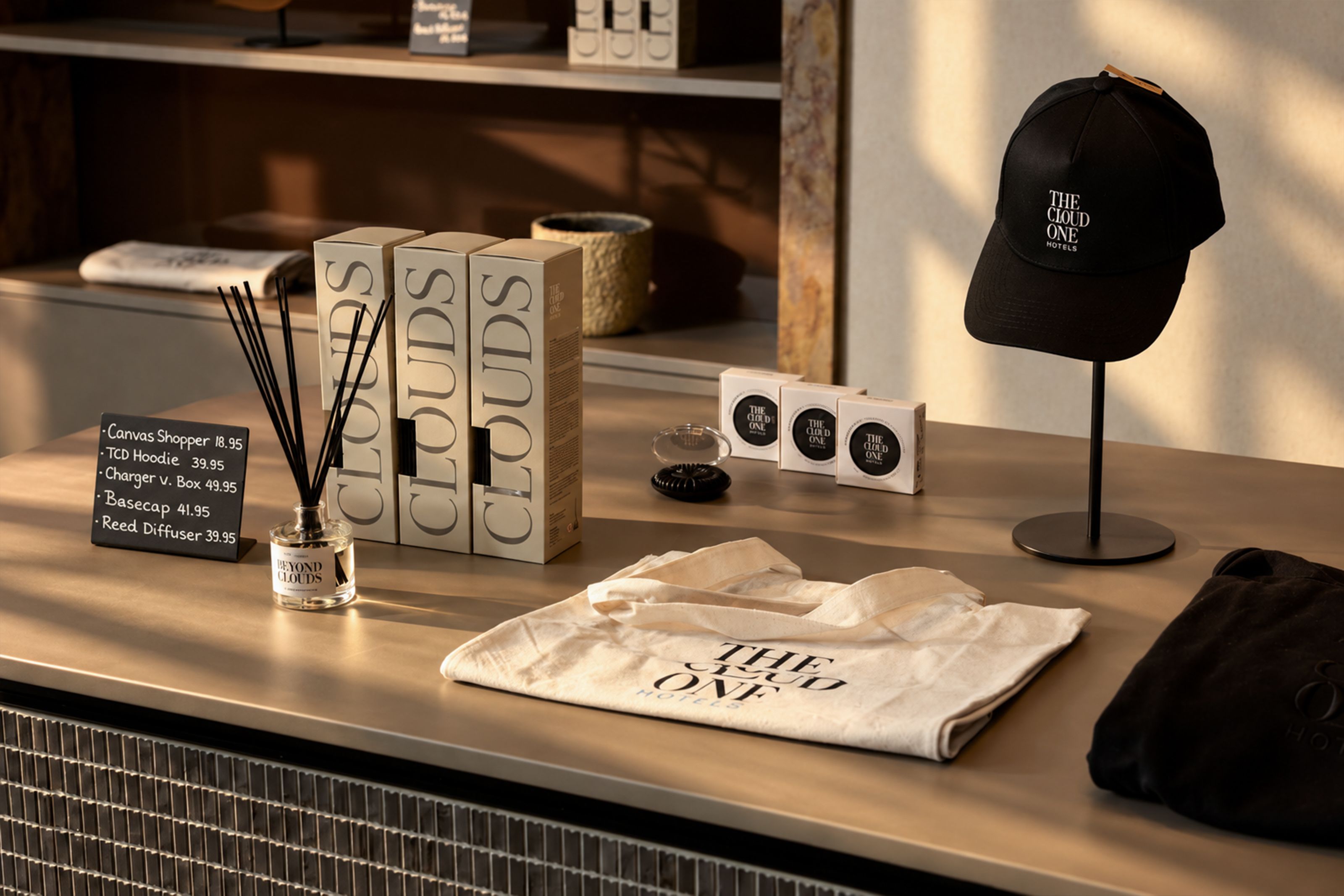 Merchandise-Artikel auf einem Verkaufstisch, darunter T-Shirts, Caps und Duftkerzen, alles mit dem Schriftzug „THE CLOUD ONE“. Die Präsentation wirkt hochwertig und stilvoll, ideal für Souvenirs oder Geschenke.