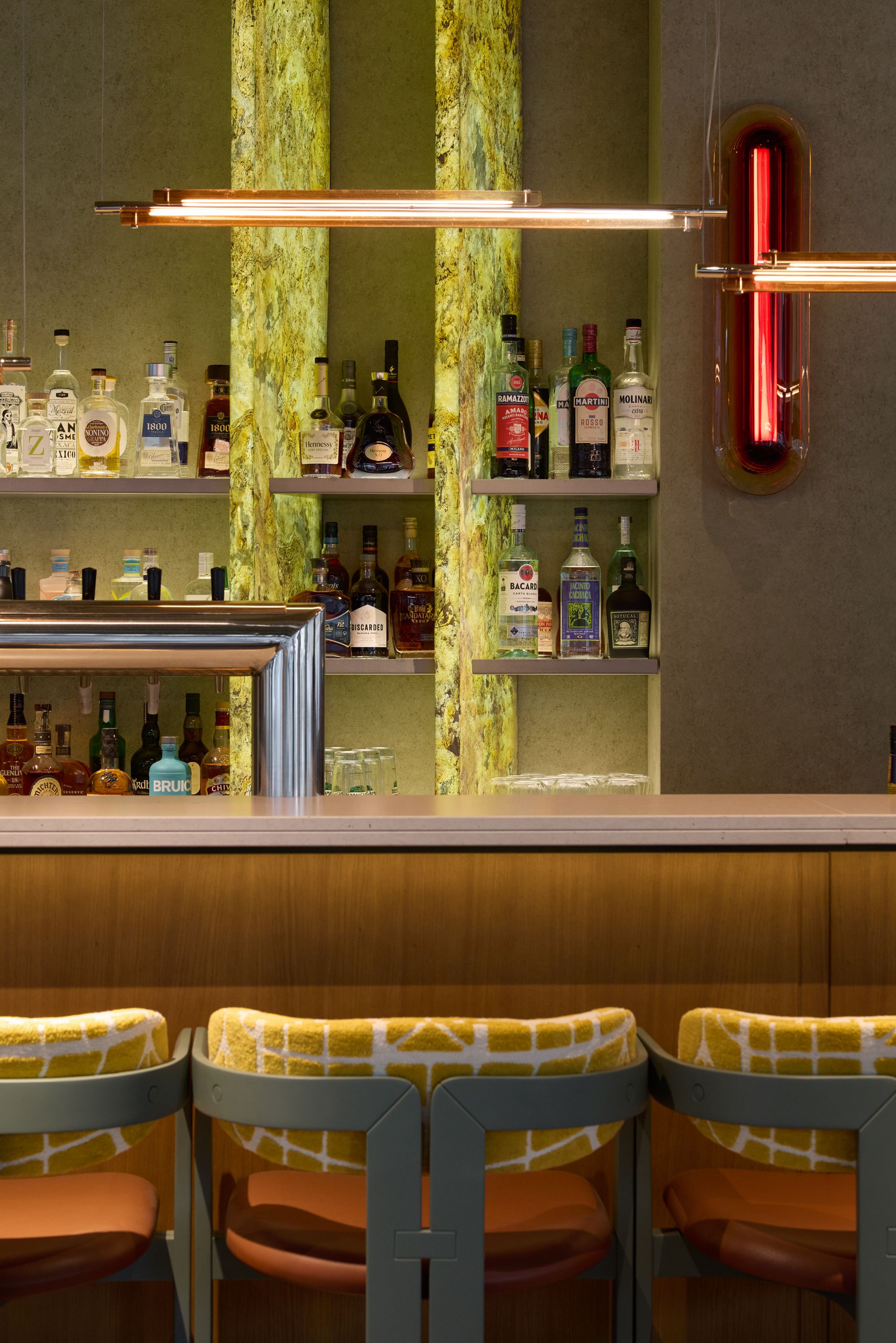 Moderne Hotelbar mit beleuchteten Regalen voller Spirituosen und stilvollen Barhockern mit gelb-weiß gemustertem Polster, geschaffen für entspannte Abende in elegantem Ambiente.