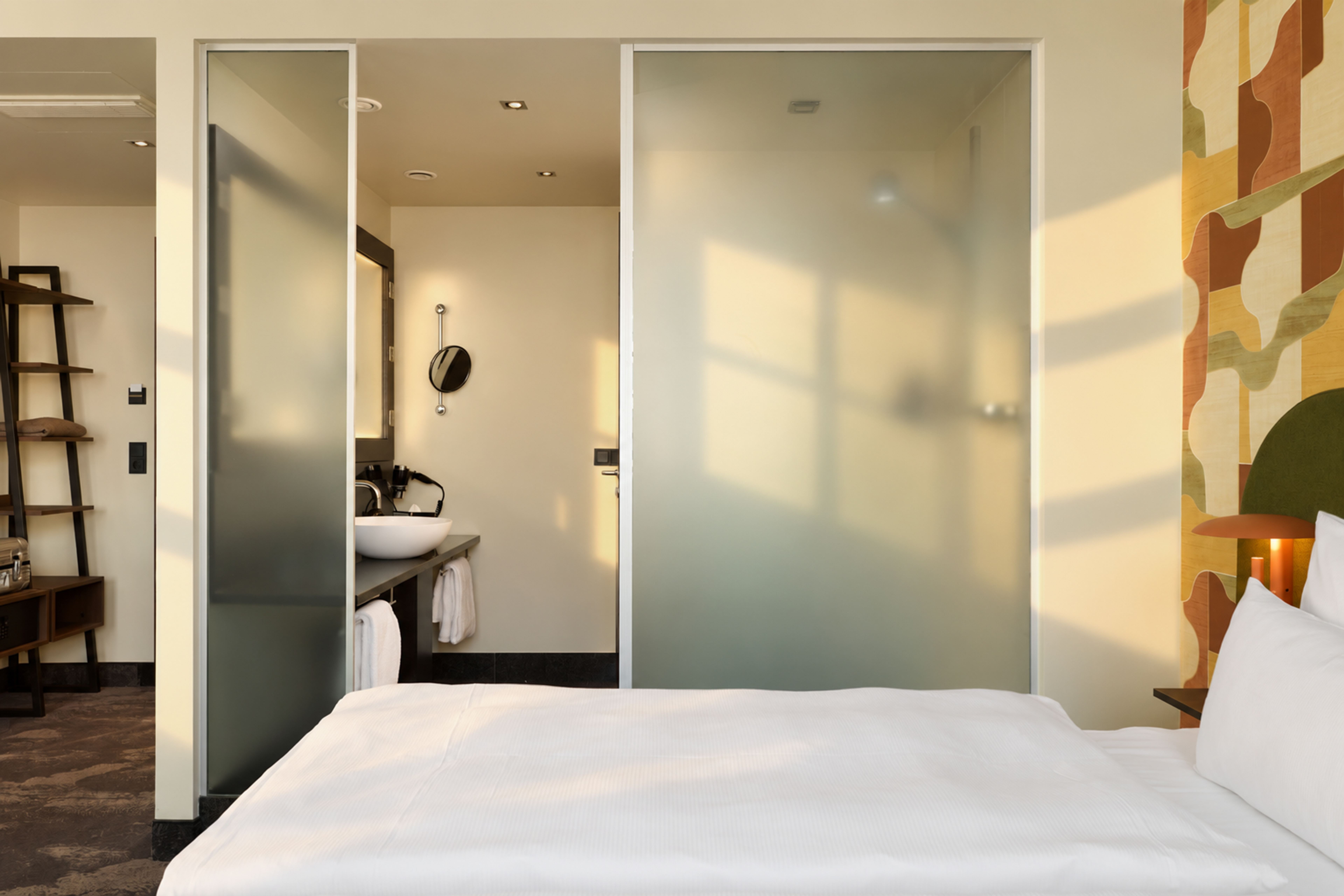 Helles Hotelzimmer mit gemütlichem Doppelbett, Tageslicht durch Fenster, halbtransparenten Glastüren zur separaten Badezimmer- und Duschzone, moderner Einrichtung und warmem, einladendem Ambiente. Helles Hotelzimmer mit gemütlichem Doppelbett, Tageslicht durch Fenster, halbtransparenten Glastüren zur separaten Badezimmer- und Duschzone, moderner Einrichtung und warmem, einladendem Ambiente.