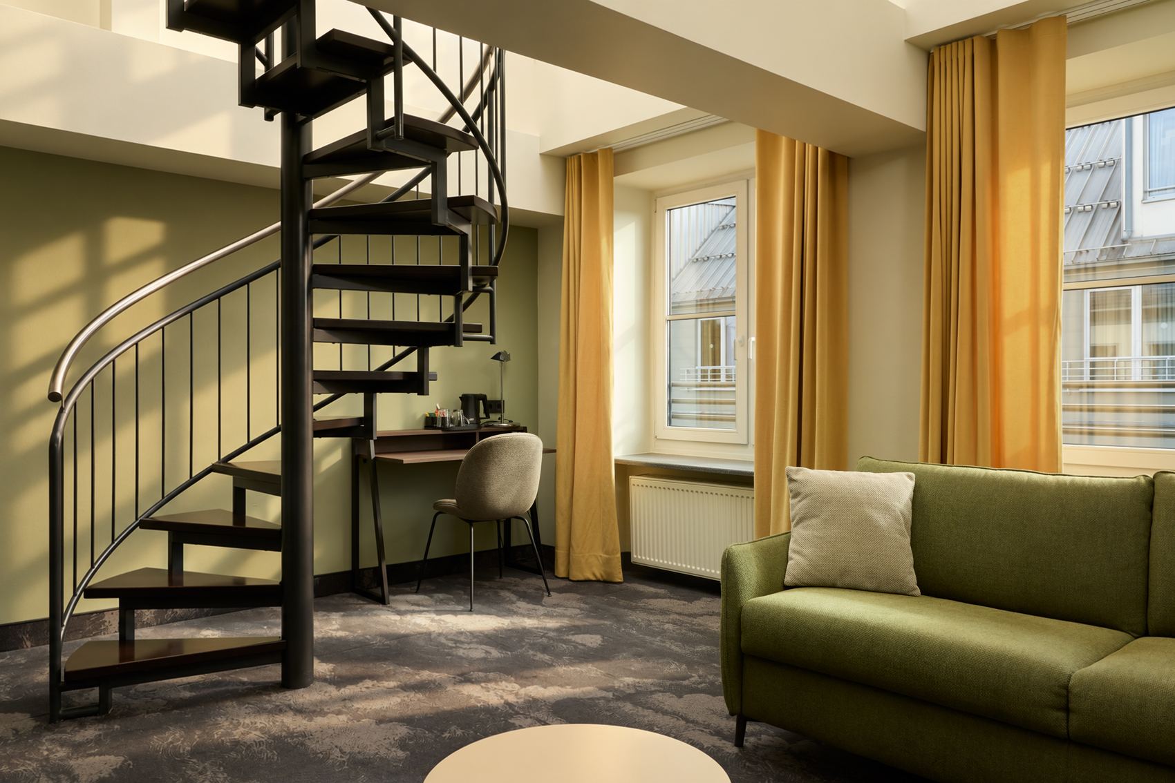 Moderner Hotelzimmerbereich mit grüner Couch, runden Couchtisch, Arbeitsplatz am Fenster und einer schwarzen Wendeltreppe zu einem oberen Bereich. Große Fenster mit gelben Vorhängen sorgen für helles, freundliches Ambiente.
