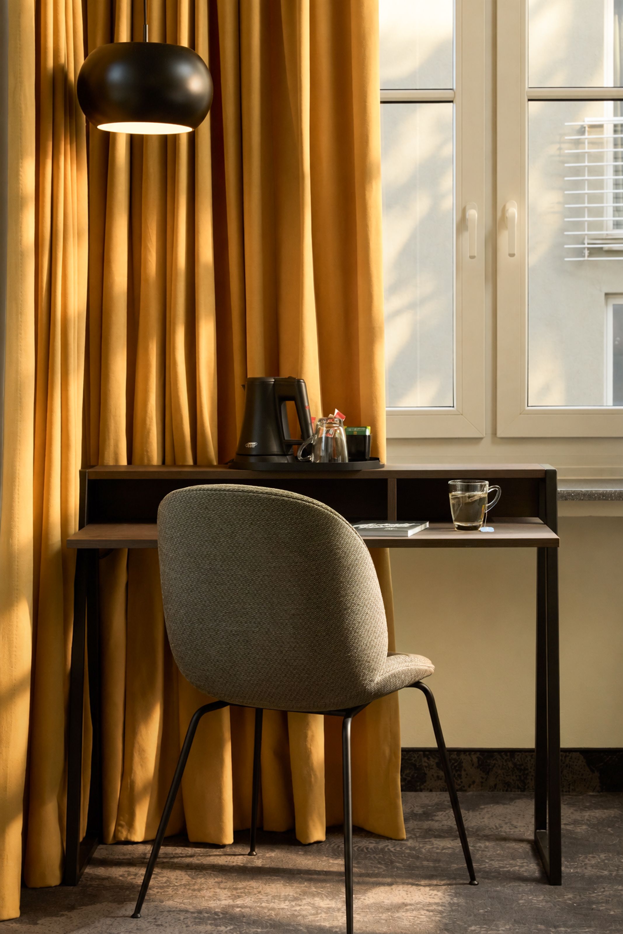 Schreibtisch mit grauem Stuhl vor großen Fenstern, warme goldgelbe Vorhänge, Kaffee- und Teezubehör auf dem Tisch, ruhige und helle Arbeits- oder Entspannungsecke im Hotelzimmer.