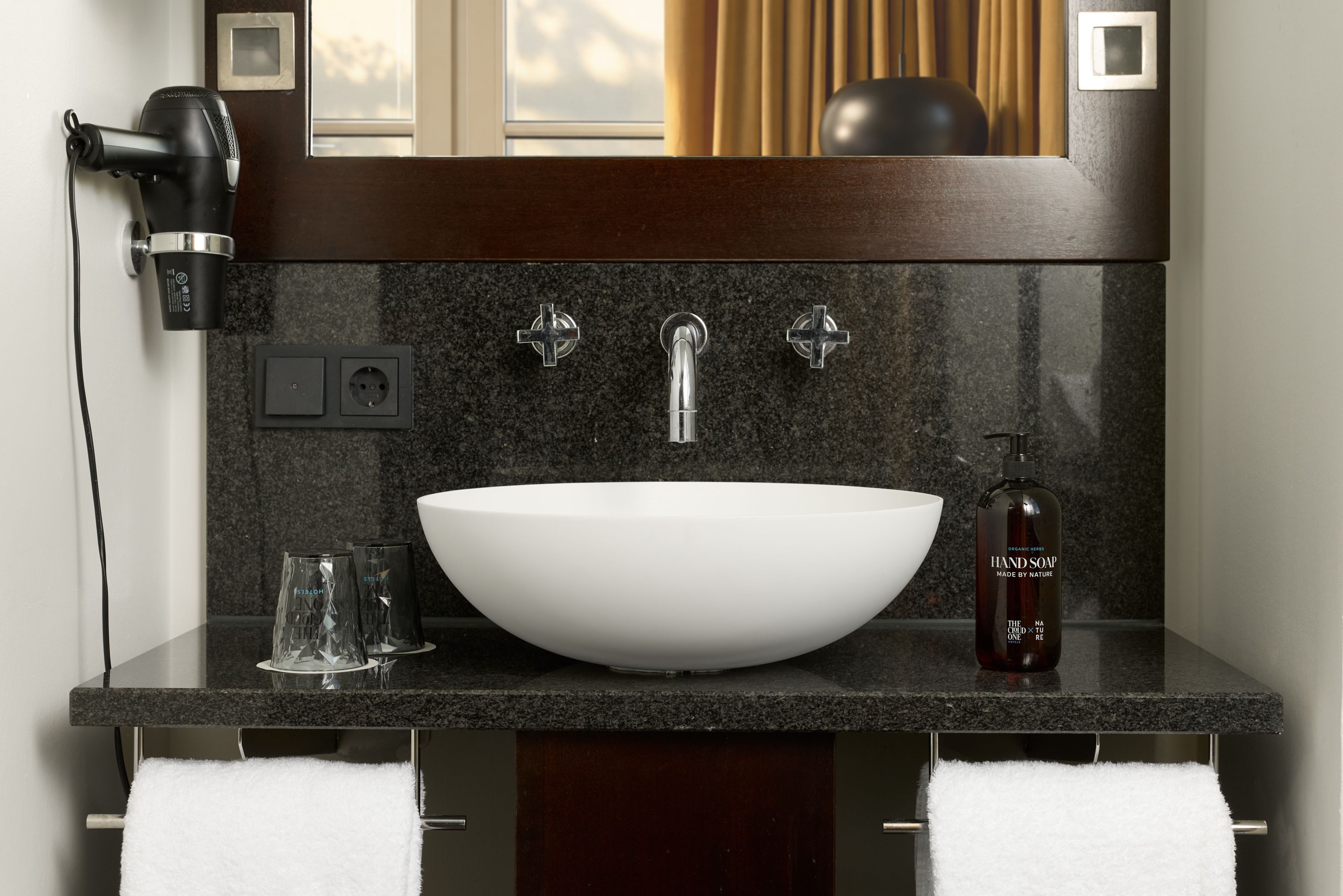 Moderne Badezimmerarmatur mit rundem, weißem Aufsatzwaschbecken auf schwarzer Arbeitsfläche. Zwei Handtücher hängen darunter, Haartrockner an der Wand, Seifenspender und zwei Gläser stehen neben dem Becken. Helle Vorhänge und Fenster im Hintergrund schaffen eine freundliche Atmosphäre.