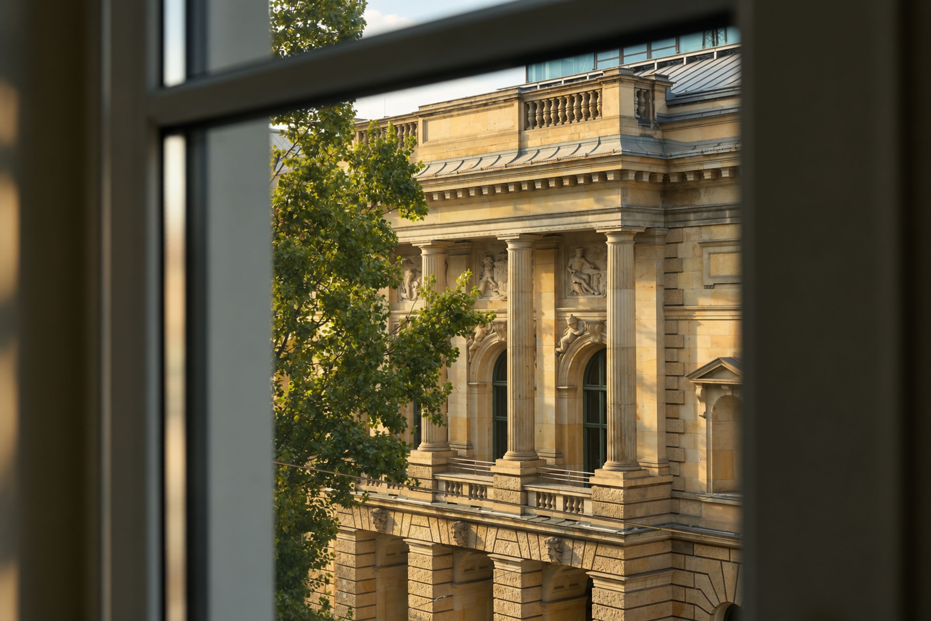 Blick aus einem Fenster auf eine historische Fassade mit Säulen und Verzierungen, umrahmt von grünen Baumzweigen, bei warmem Tageslicht.