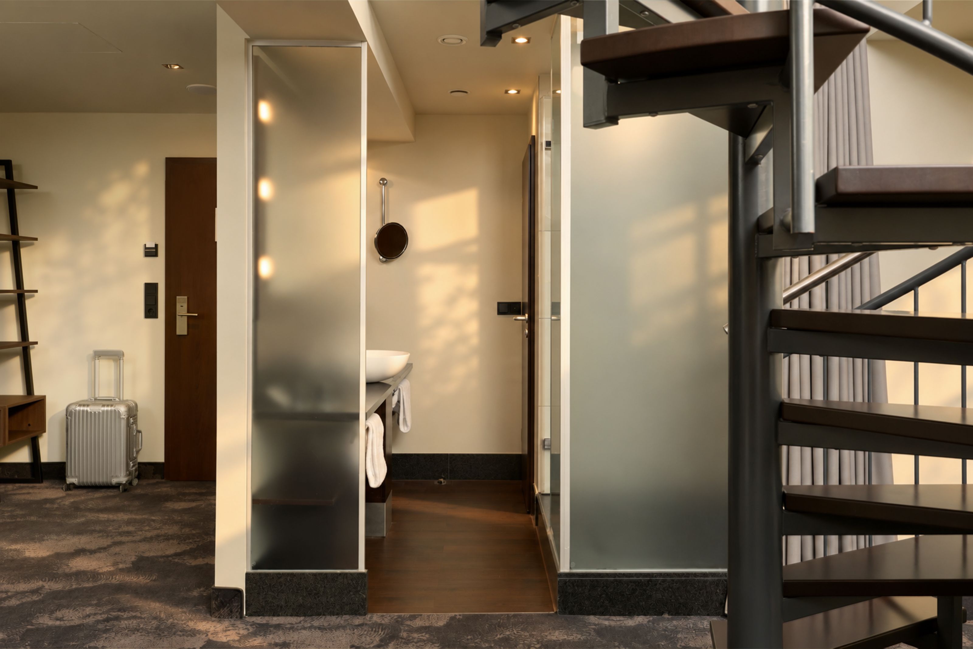 Helles Hotelzimmer mit offenem Badbereich, Duschkabine und moderner Wendeltreppe. Sonnenlicht fällt durch die Fenster und erzeugt warme Atmosphäre. Koffer steht im Raum, der Boden ist mit Teppich ausgelegt.