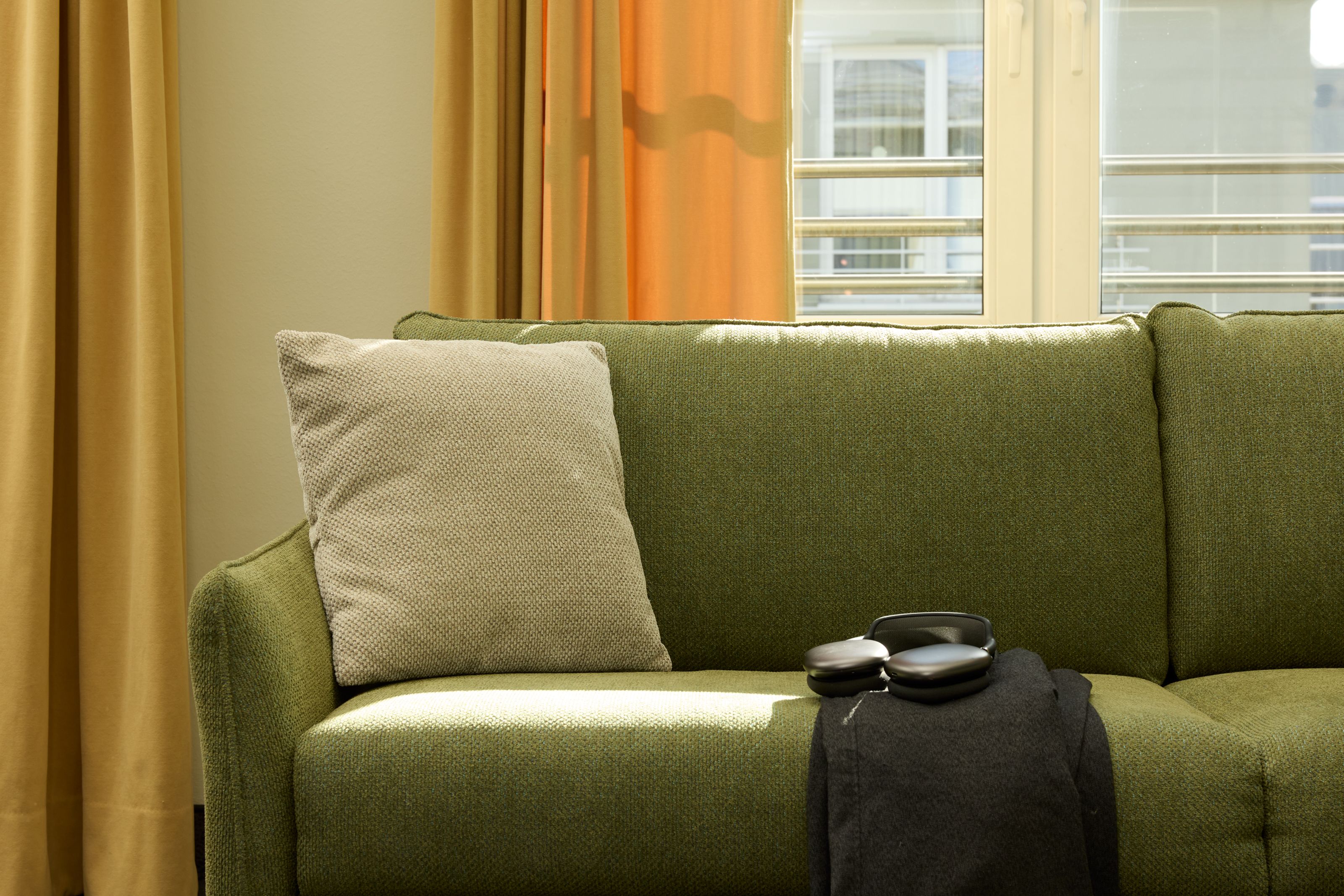Grünes Sofa mit einem grauen Kissen, auf dem eine schwarze Jacke und Kopfhörer liegen. Helles Tageslicht fällt durch ein Fenster mit gelben Vorhängen.