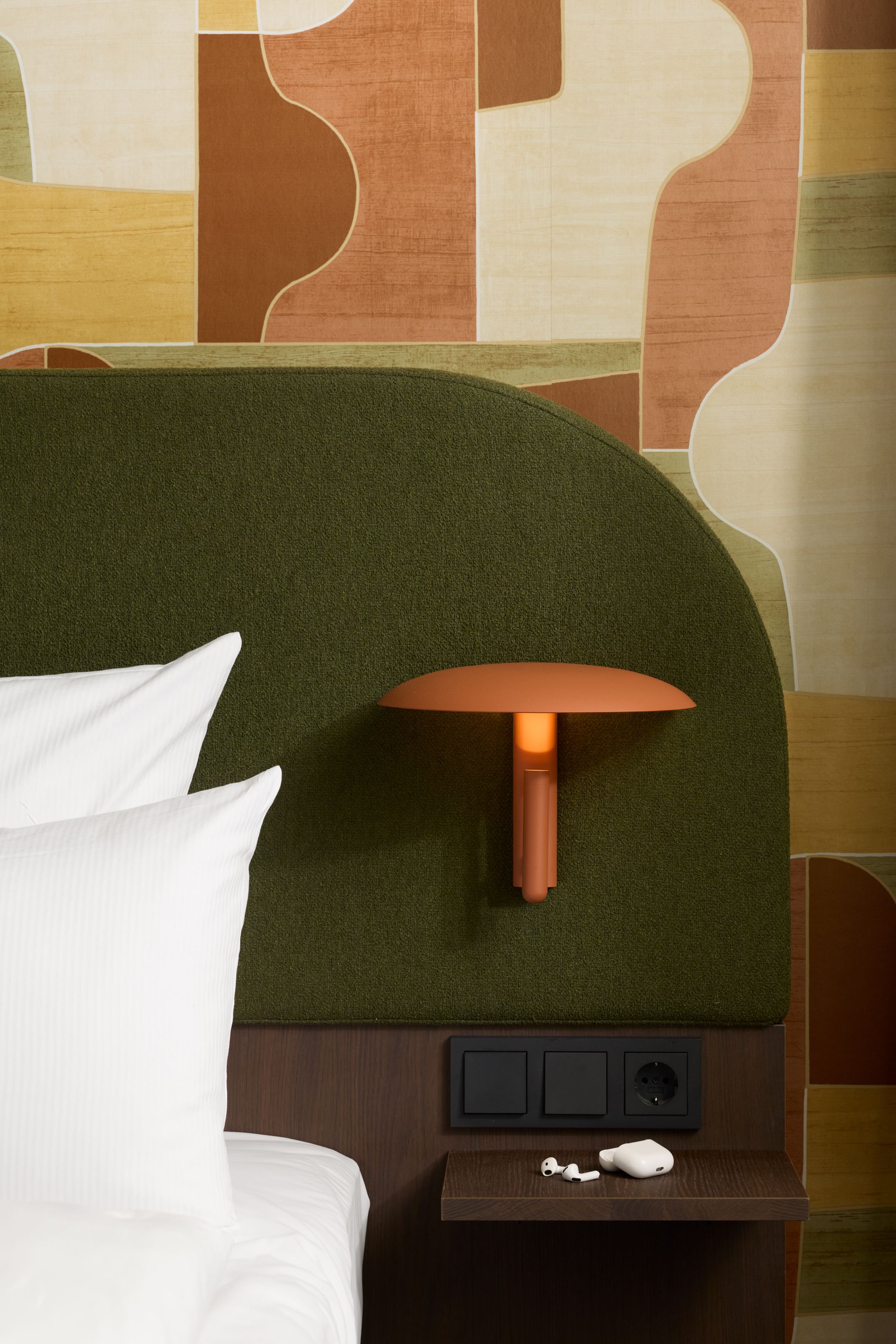 Bequeme Hotelbett-Ecke mit grünem gepolstertem Kopfteil, modernem orangeroten Wandlicht und Wand mit abstraktem retrofarbigem Muster, neben kleinem Nachttisch mit Steckdosen und weißen Kopfhörern.