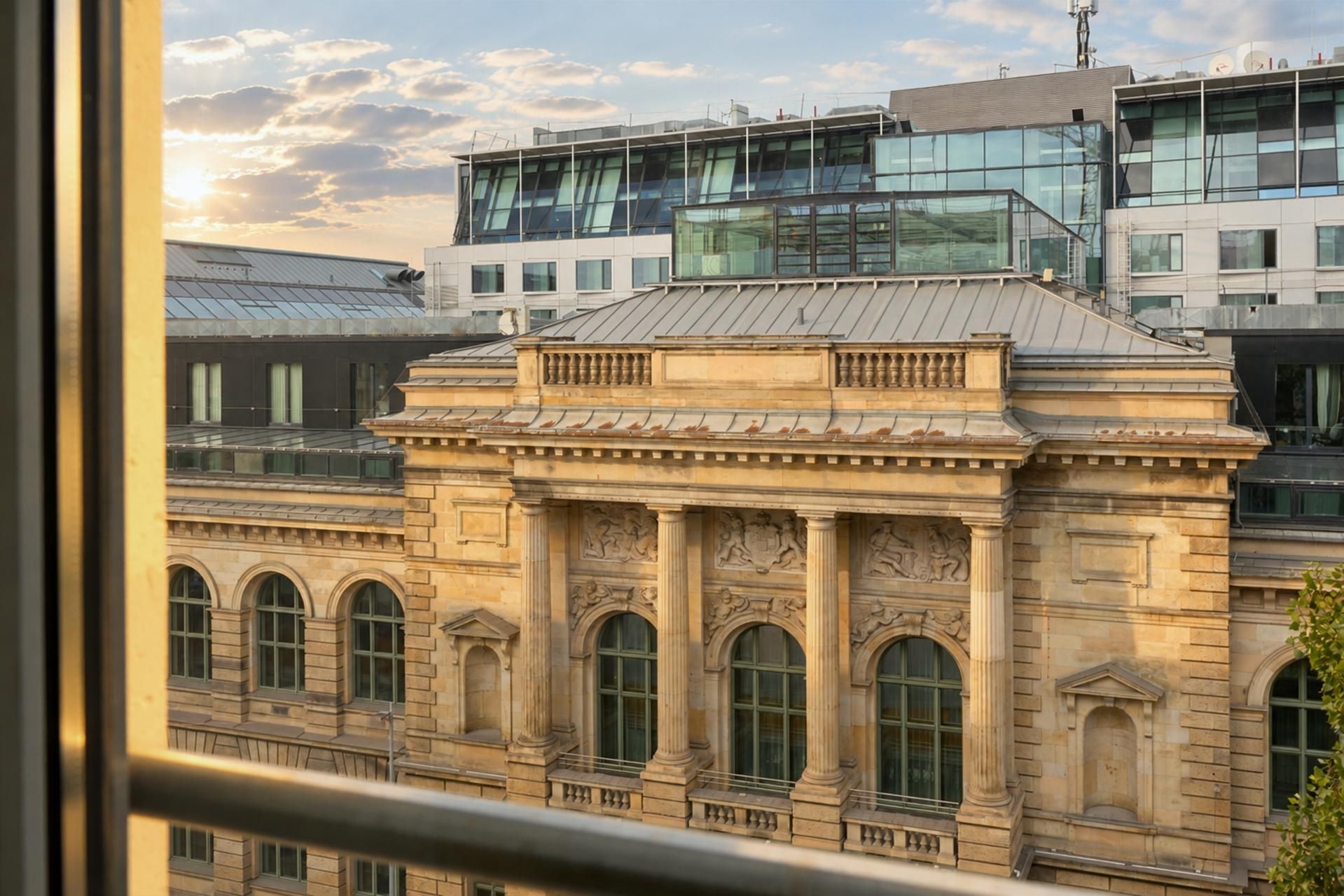 Blick aus einem Fenster auf ein historisches Sandsteingebäude mit hohen, verzierten Fenstern, dahinter moderne Bürogebäude unter einem sonnigen Himmel mit vereinzelten Wolken.