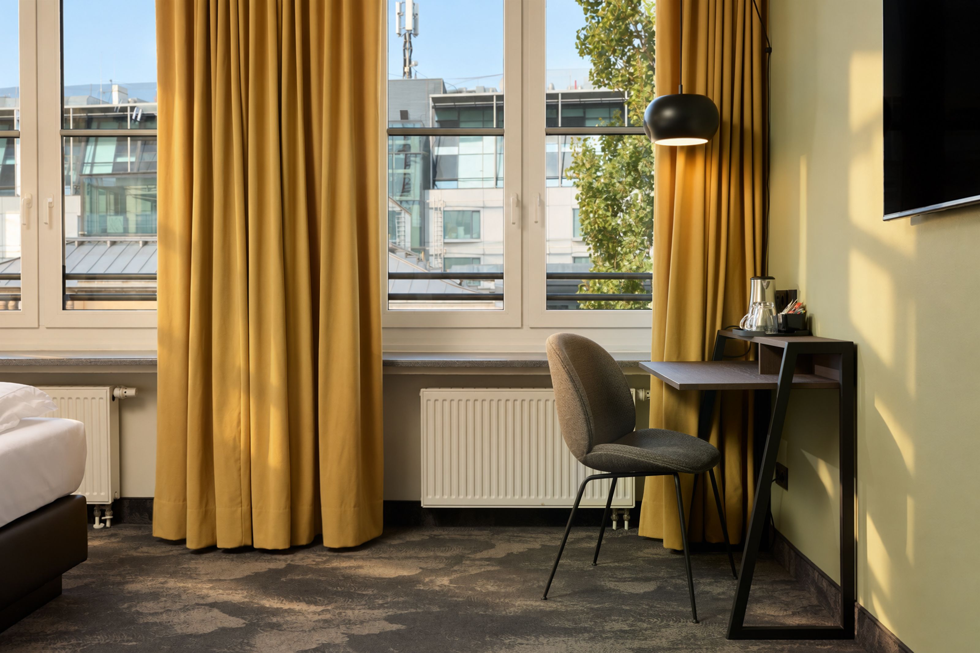 Helles Hotelzimmer mit großen Fenstern, gelben Vorhängen und modernem Arbeitsplatz mit Stuhl, Schreibtischlampe und Fernseher an der Wand, gemütliche Atmosphäre für Arbeit und Entspannung.