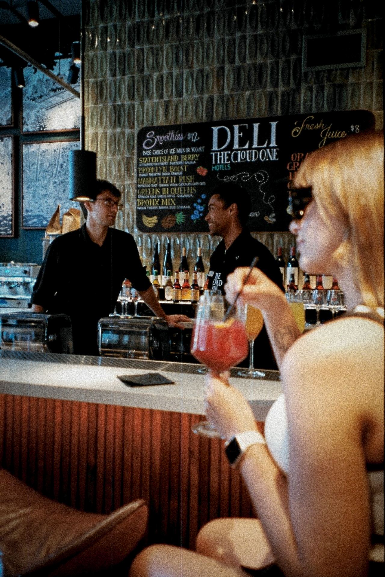 Frau mit Cocktail sitzt an einer Hotelbar, zwei Barkeeper unterhalten sich hinter dem Tresen, moderne Atmosphäre, Getränkeauswahl im Hintergrund.