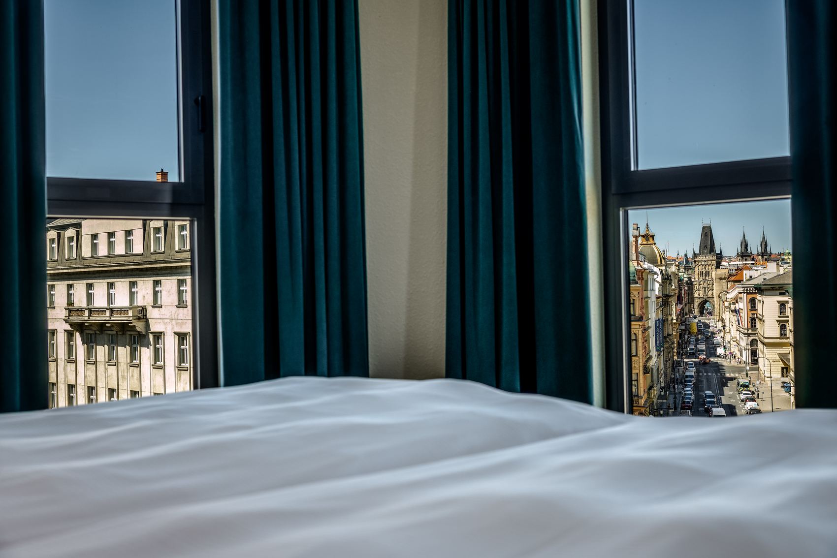 Blick aus einem Hotelzimmer mit weißer Bettdecke durch zwei Fenster auf eine belebte Stadtstraße und historische Gebäude bei Tageslicht.