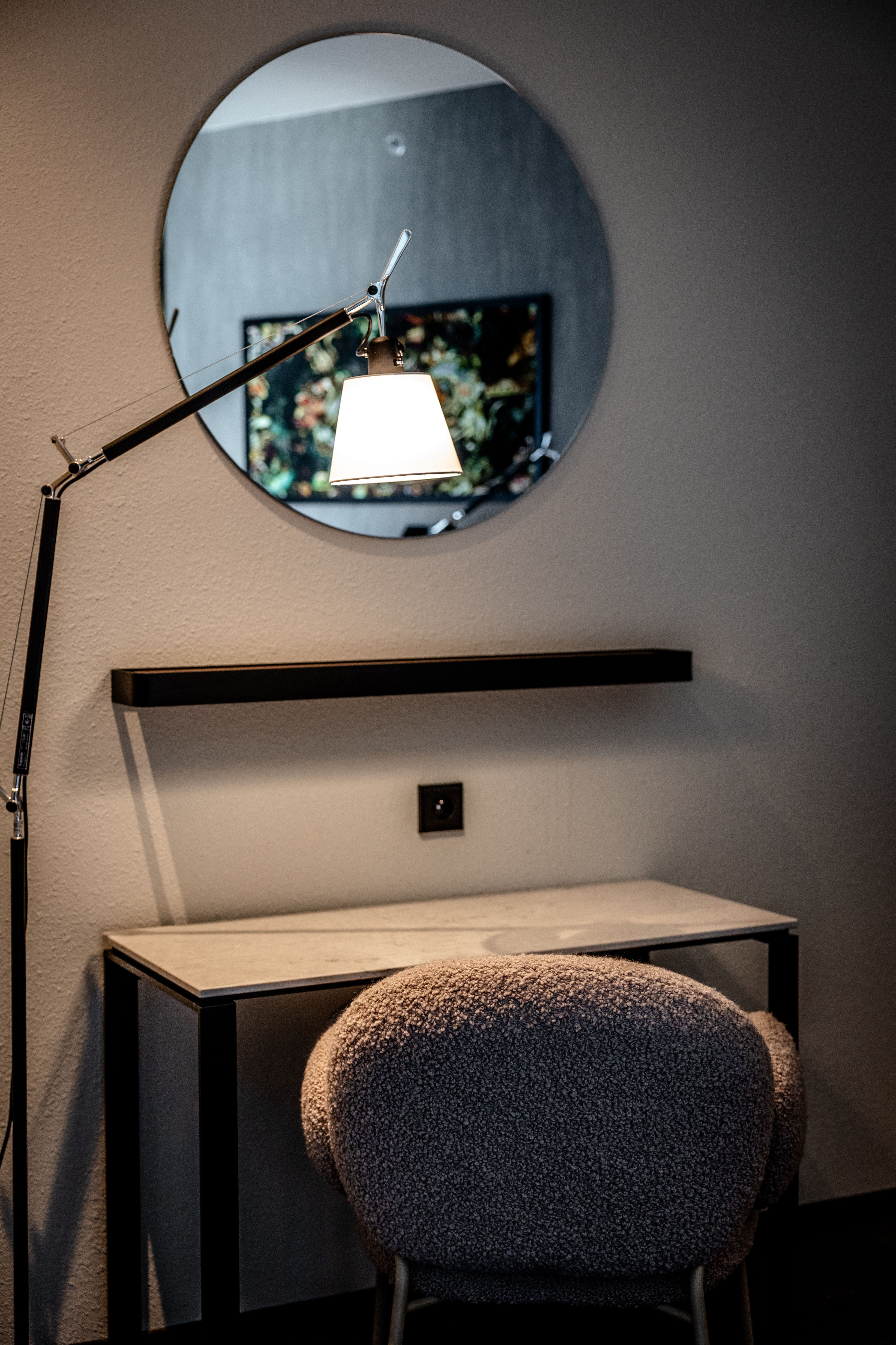 Runder Spiegel über einem schlichten Schreibtisch mit grauem, weichem Stuhl, beleuchtet von einer verstellbaren Wandlampe, die warmes Licht auf die Oberfläche wirft.