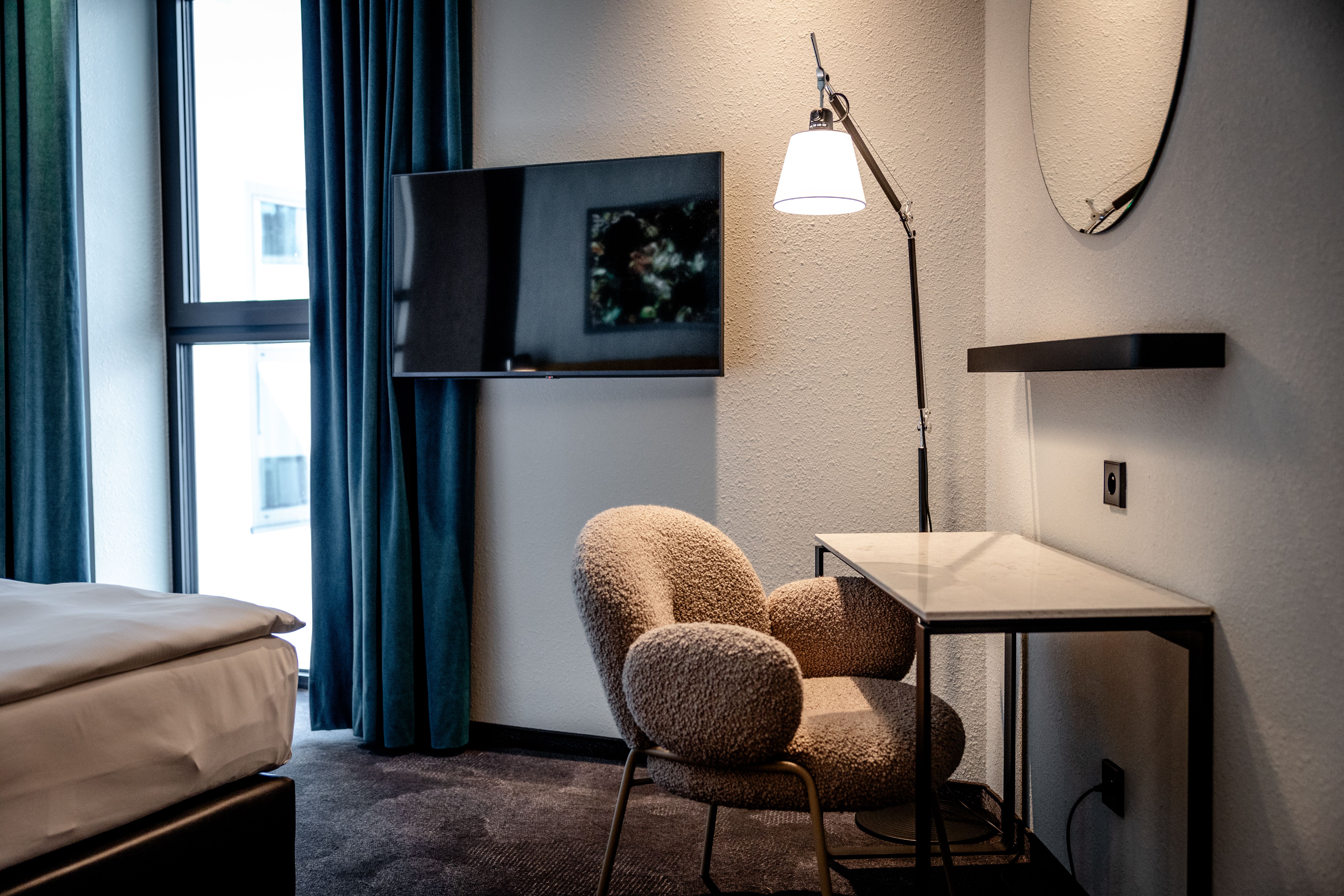 Modernes Hotelzimmer mit gemütlichem Sessel, Schreibtisch mit Lampe, Wandspiegel und Flachbildfernseher, neben einem großen Fenster mit dunklen Vorhängen.