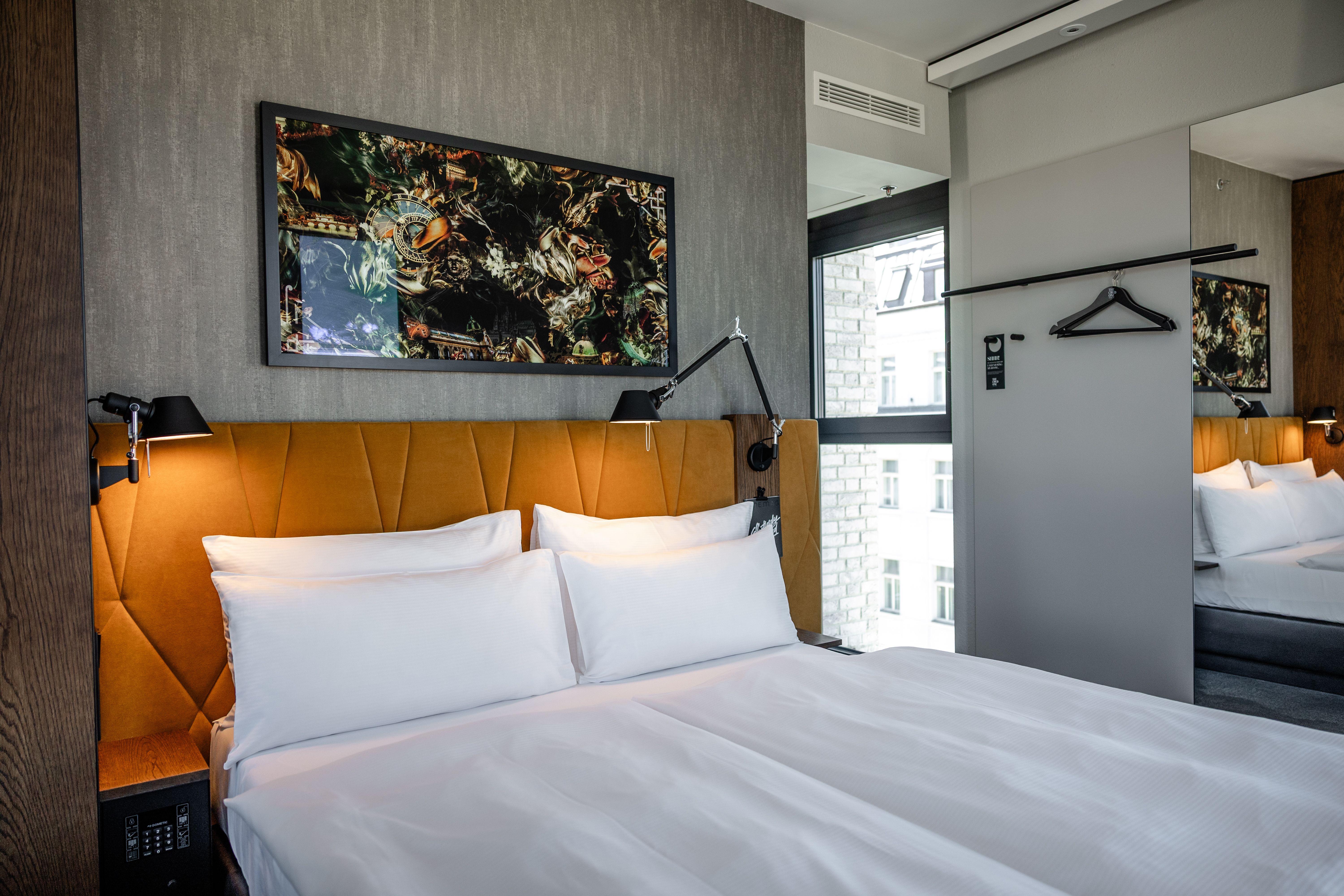 Modernes Hotelzimmer mit gemütlichem Doppelbett, hellen weißen Bettwaren, stilvollem gepolstertem Kopfteil in warmem Orange und dekorativem Kunstbild über dem Bett. Große Fenster bieten Tageslicht und Aussicht. Minimalistische, elegante Einrichtung für komfortablen Aufenthalt.